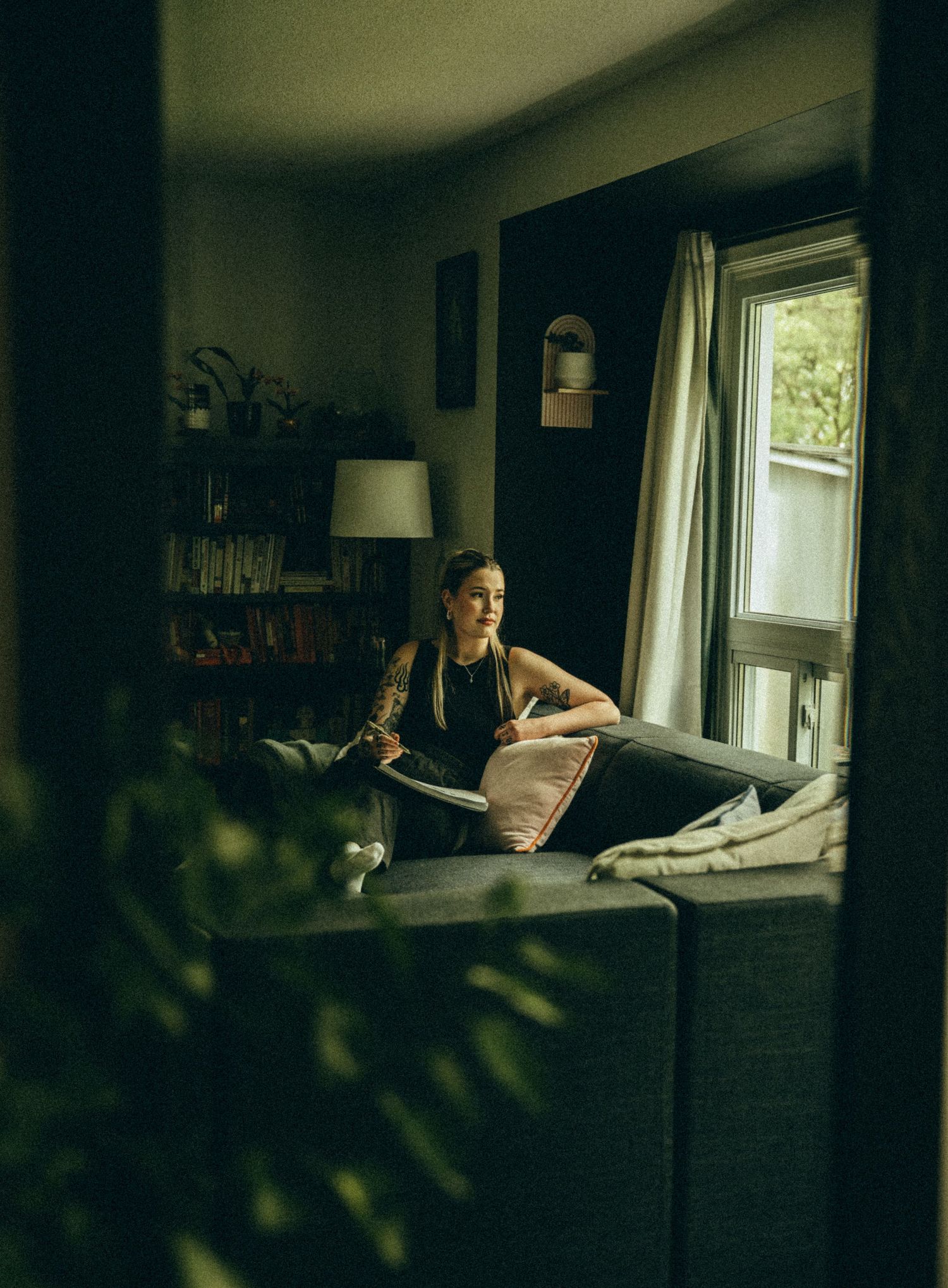 Tattooed woman relaxing on gray sofa in cozy dimly lit living room near a bright window.