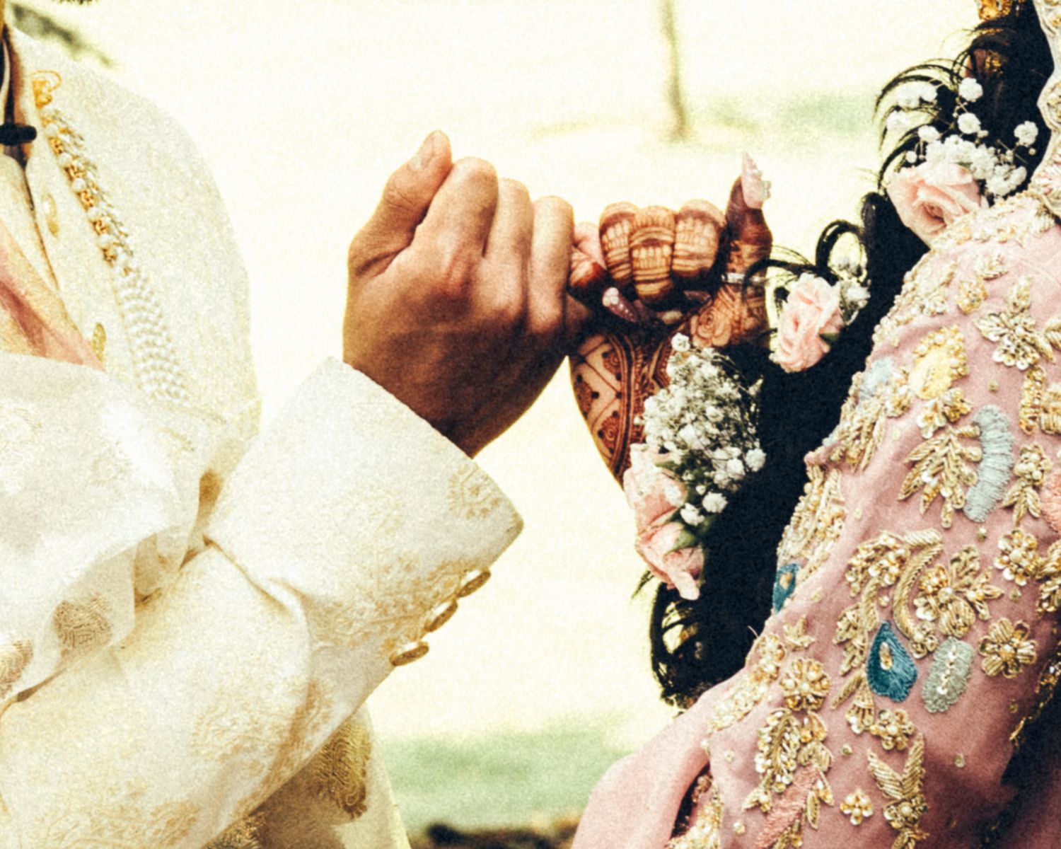 Tender moment of a groom in white sherwani holding the henna-adorned hand of his bride in a pink embroidered lehenga.