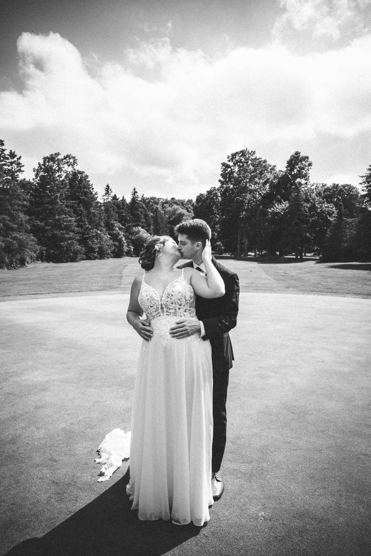 Black and white photo of bride and groom kissing on a golf course under dramatic cloudy sky.