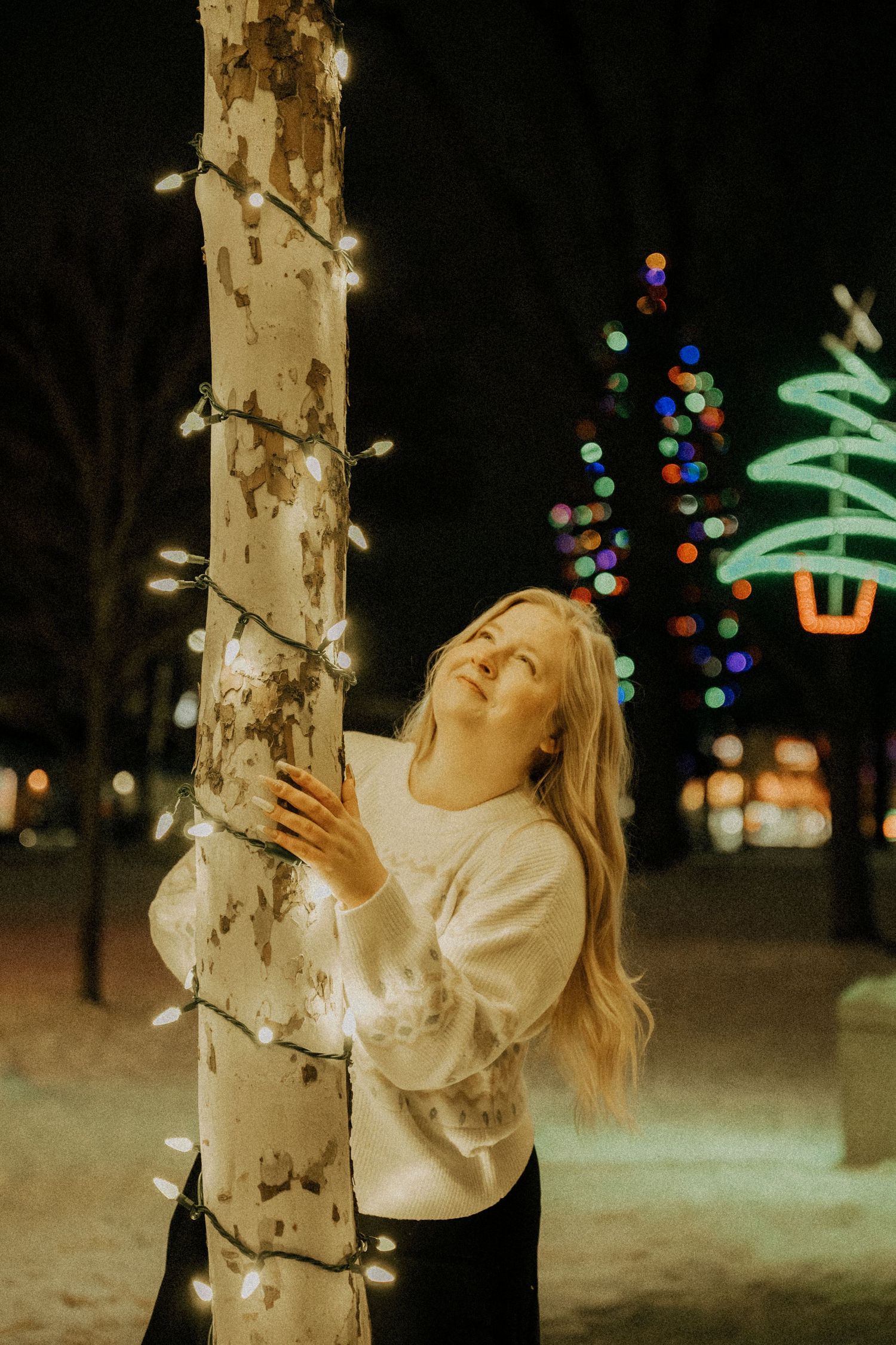 Blonde woman embracing a tree wrapped in white fairy lights at night.