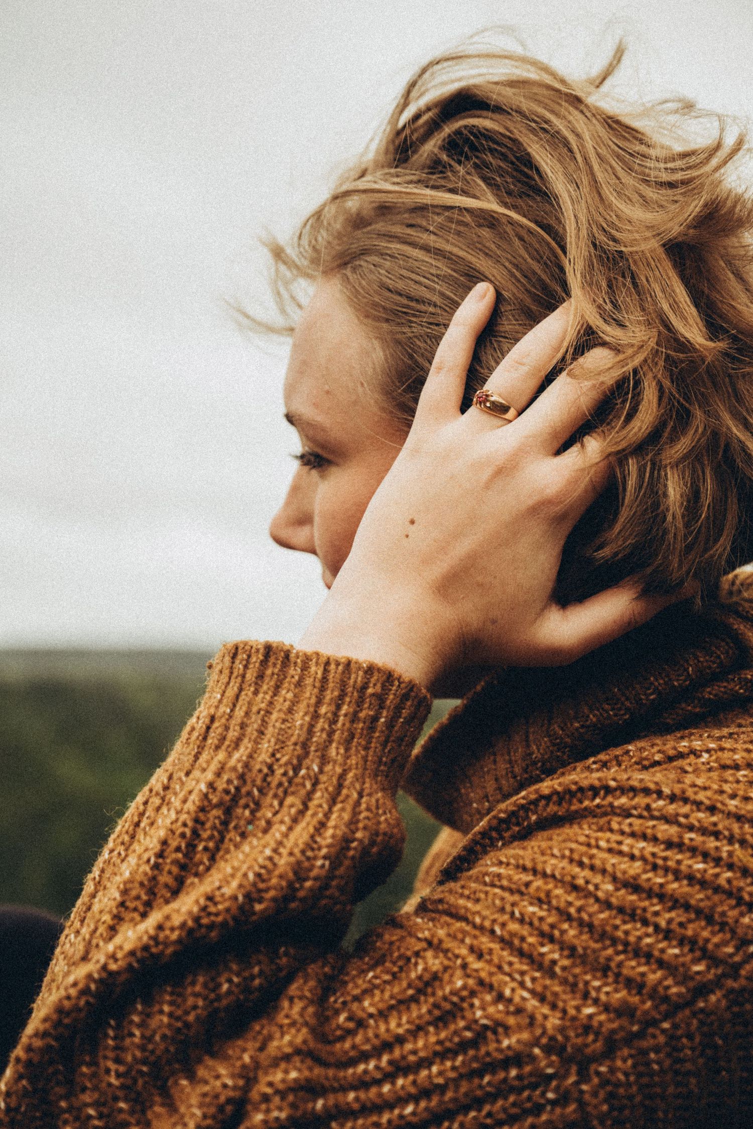 A woman in a brown knit sweater touches her windswept blonde hair outdoors.