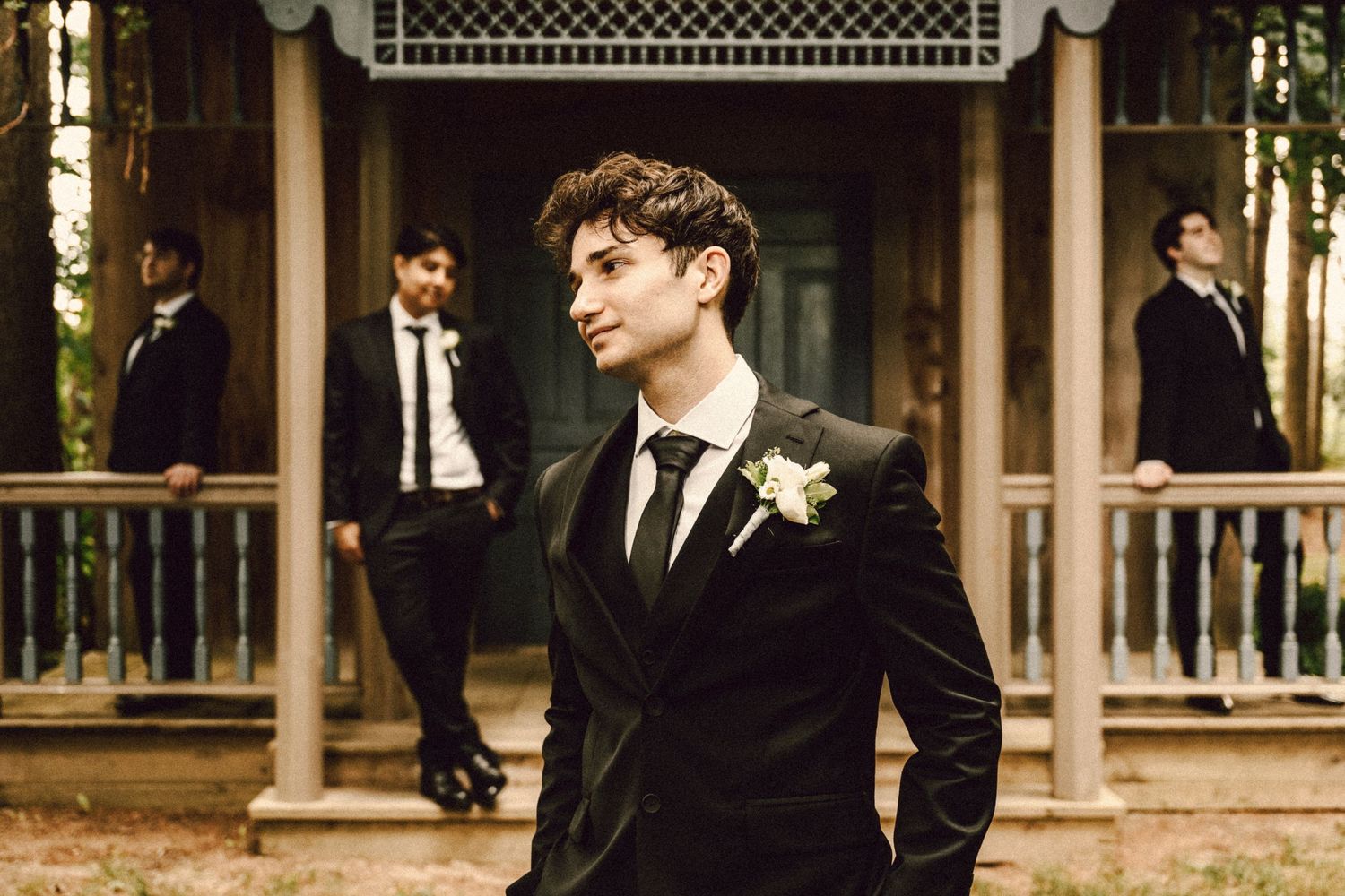 Dapper groom in black velvet suit with white boutonniere stands before rustic venue with groomsmen.