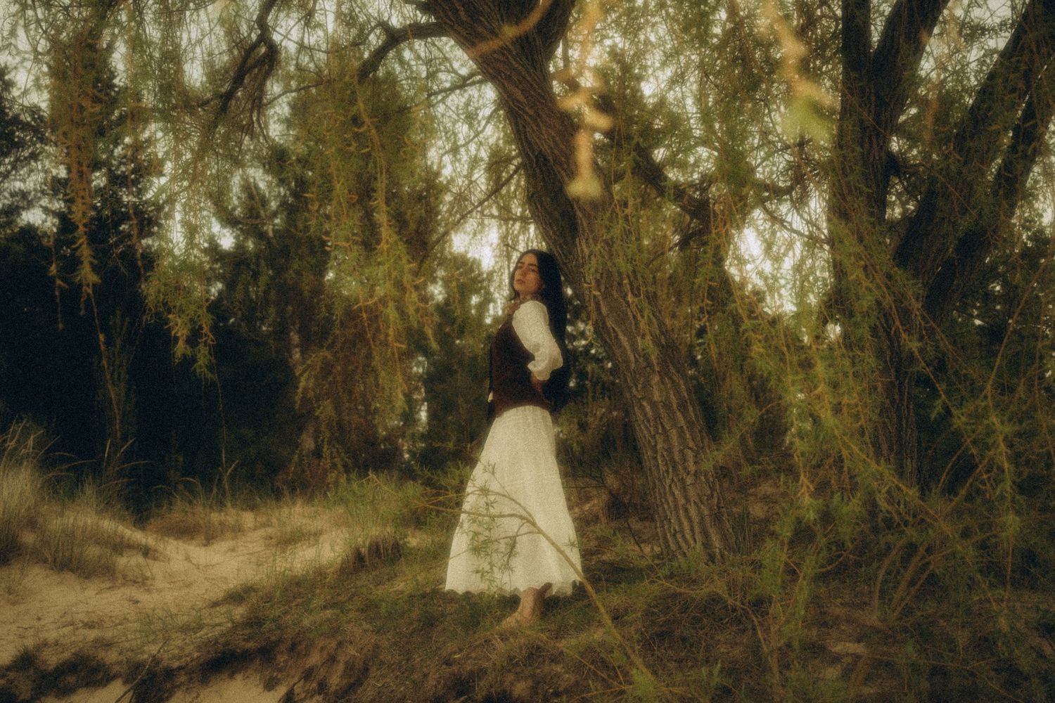 Woman in white skirt standing barefoot among lush green trees in dreamy forest setting.