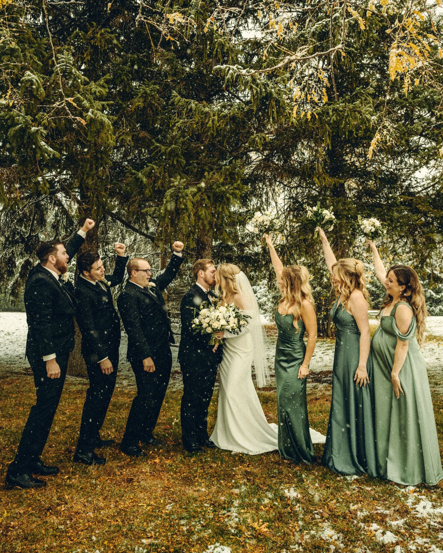 Wedding party celebrating in snow with bridesmaids in green dresses and groomsmen in black suits.