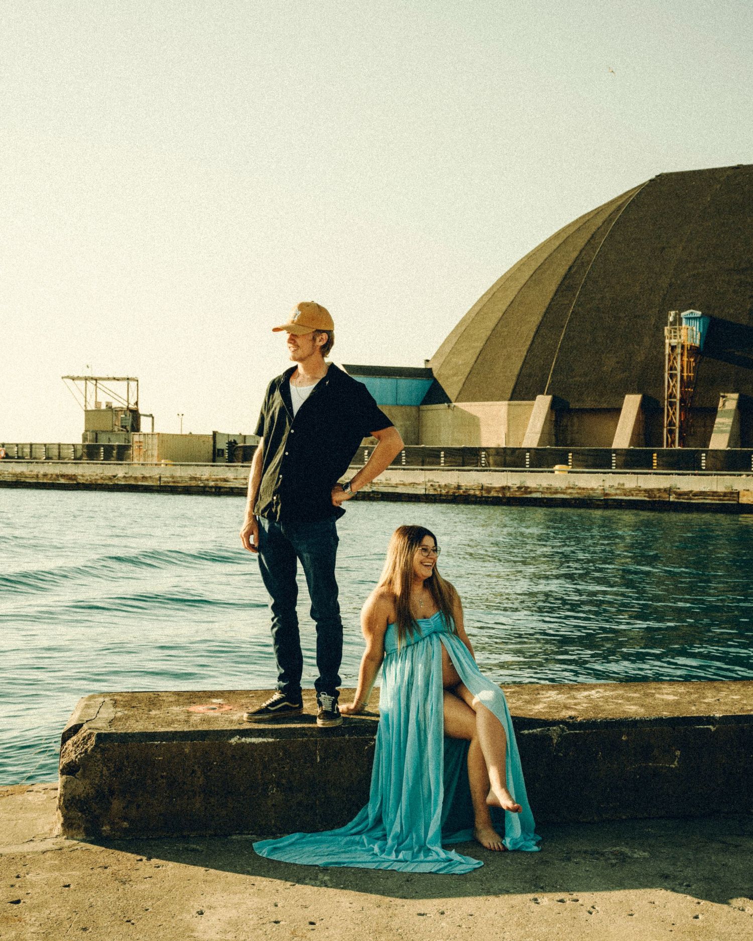Couple poses by waterfront, woman in flowing teal gown sits while man stands beside her near a domed building.