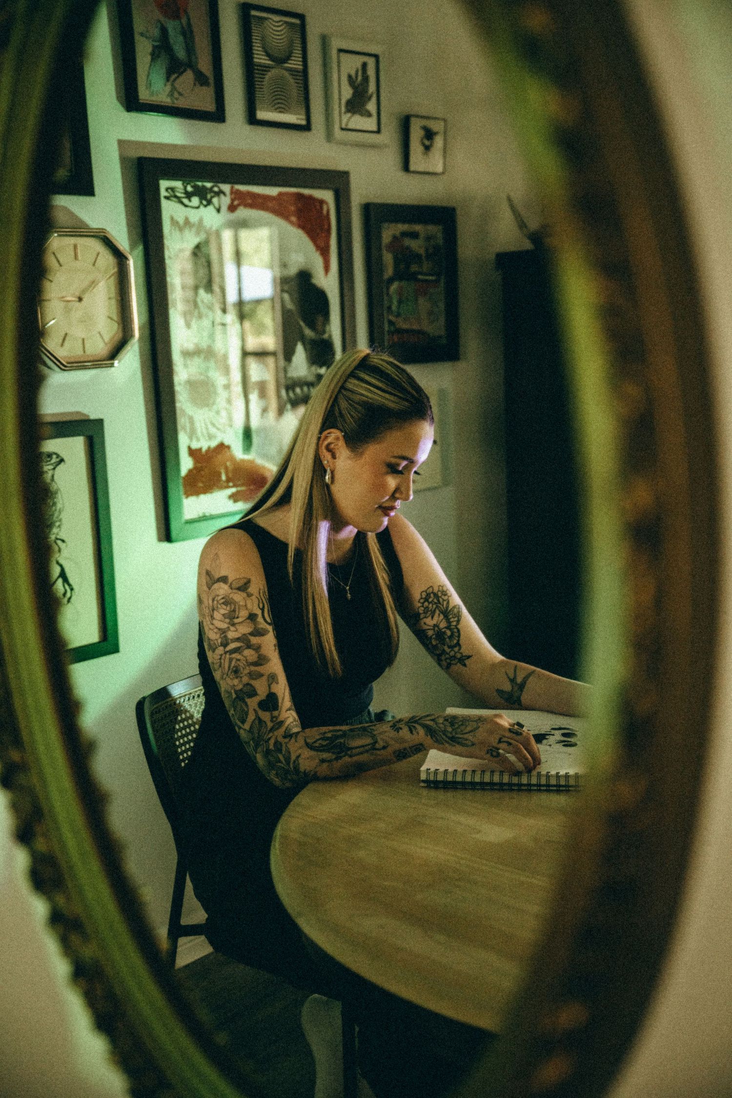 Tattooed woman sketching in notebook at wooden table, reflected in ornate gold mirror surrounded by framed artwork.