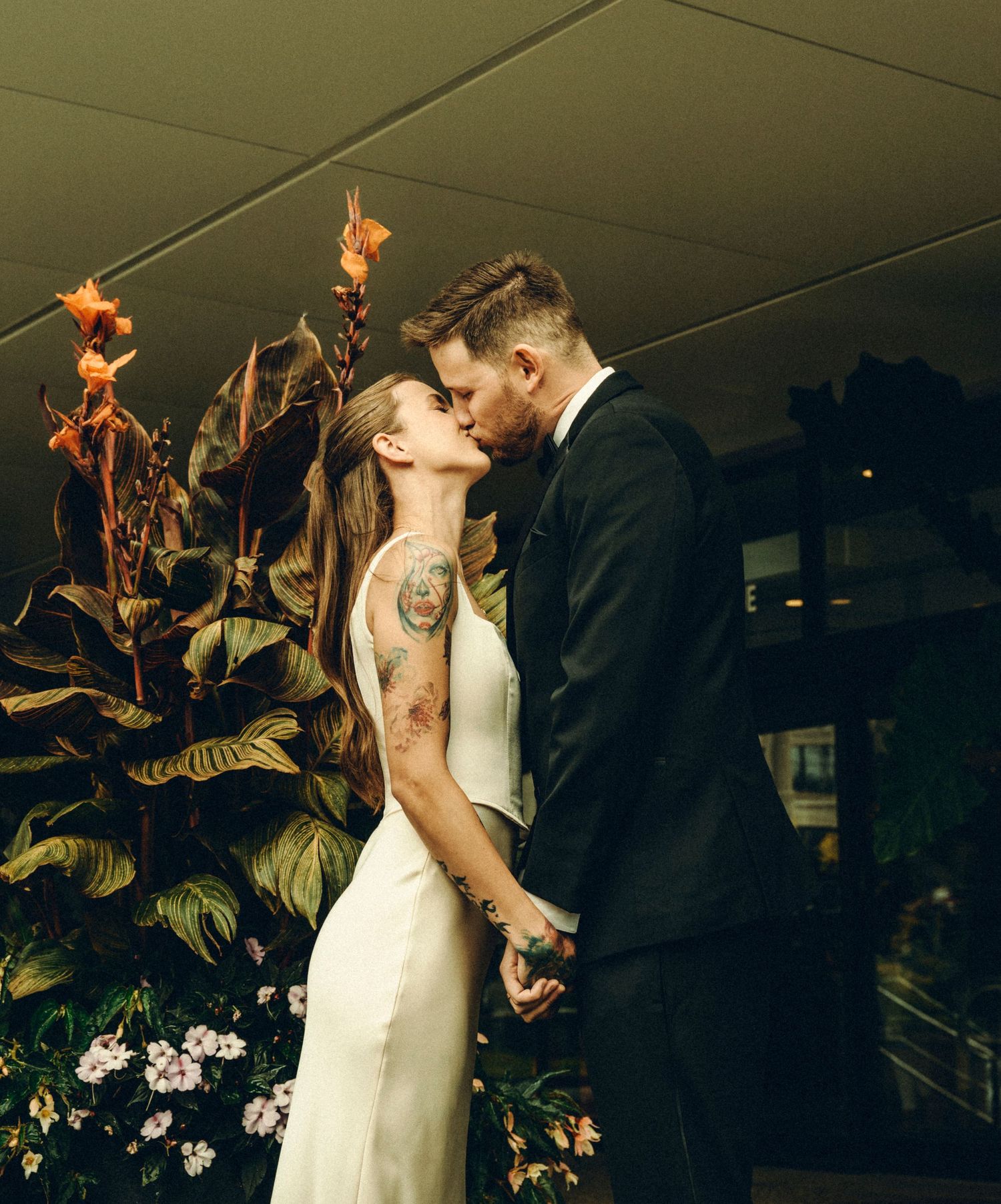 Tattooed bride and groom sharing romantic kiss beside tropical floral arrangement indoors.