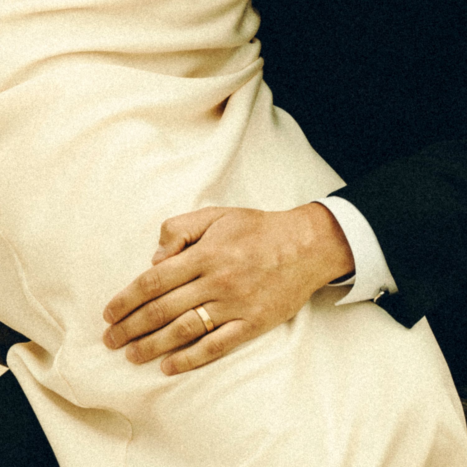 Close-up of groom's hand with gold wedding band resting on bride's white wedding dress.