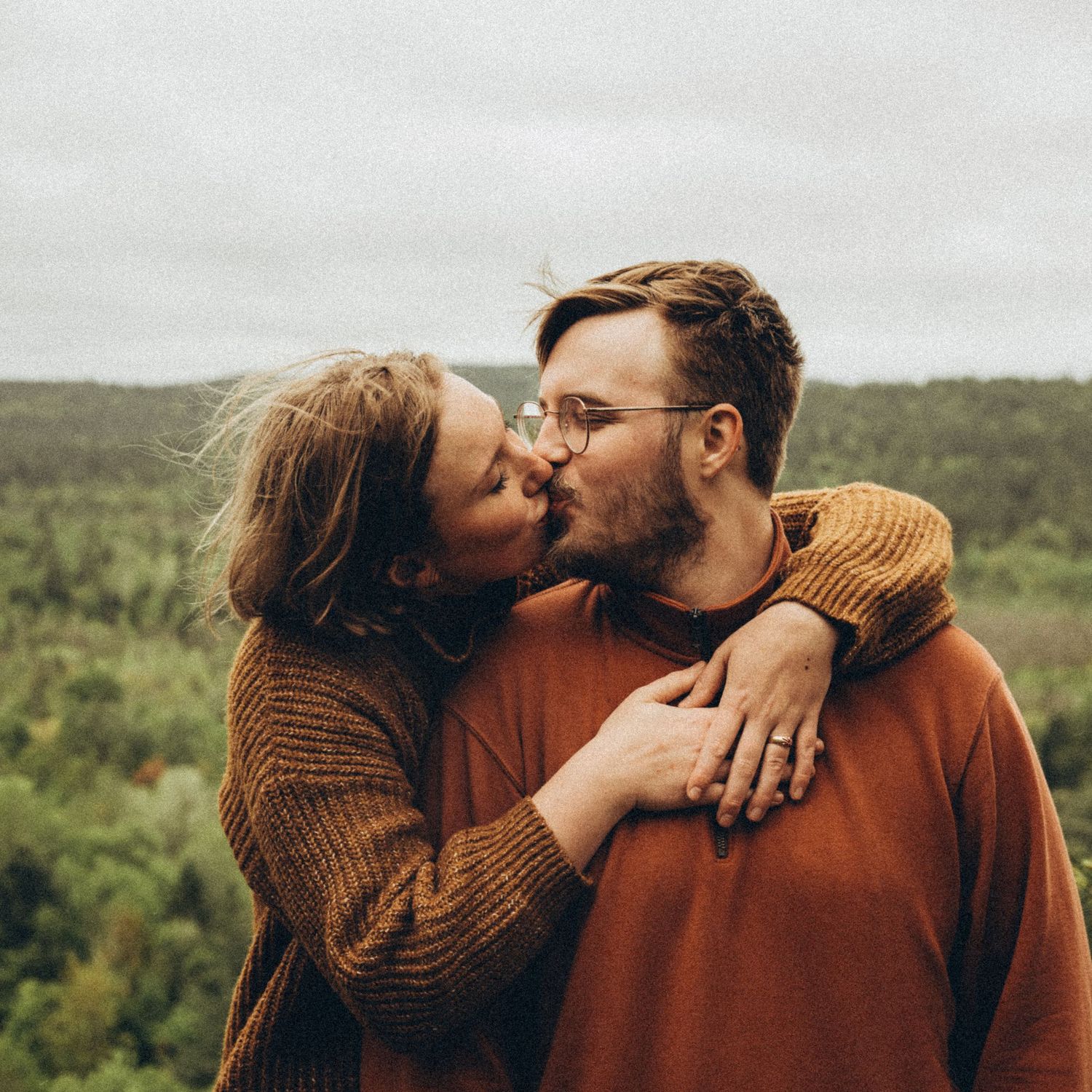 A couple sharing a kiss outdoors with a lush green forest landscape behind them.