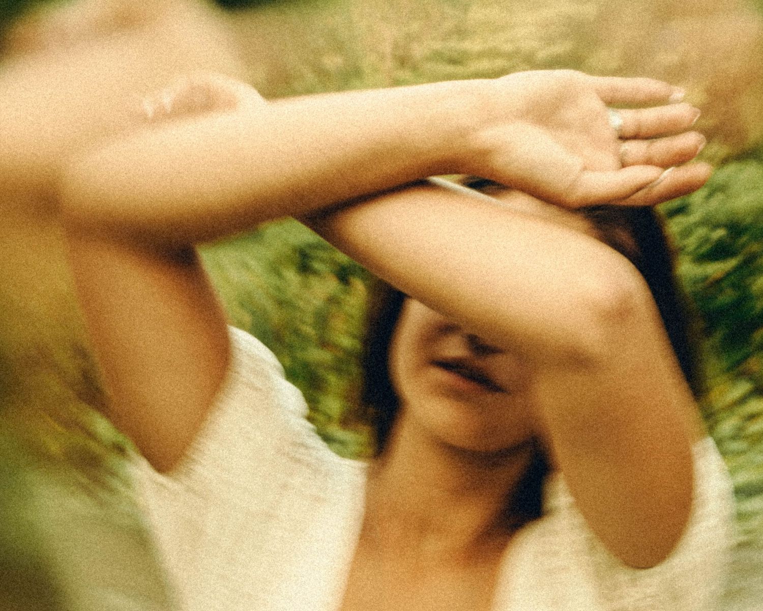 Blurred artistic photo of woman in white top with arms raised covering face outdoors.