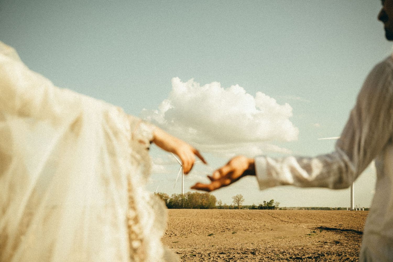 Bride and groom reaching toward each other in an open field under a bright cloudy sky.