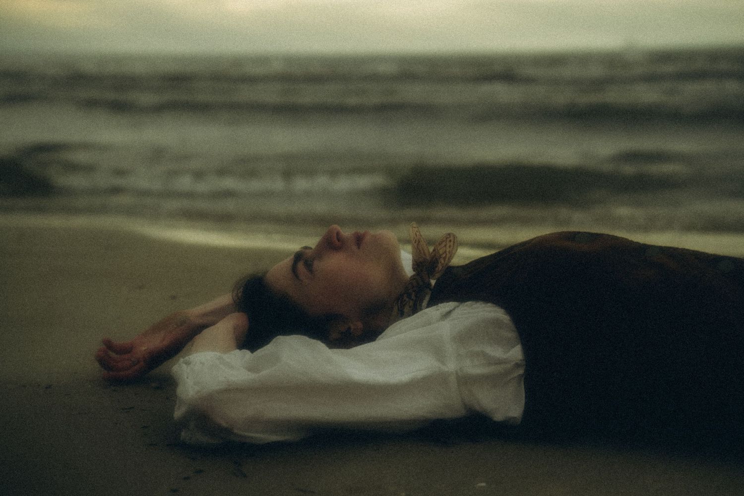Woman in white blouse lying on sandy beach with moody ocean waves in background.