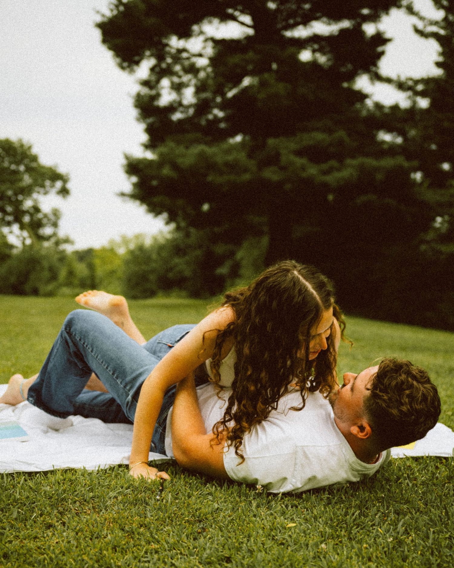 Couple kissing and laughing together while lying on white blanket on green grass outdoors.