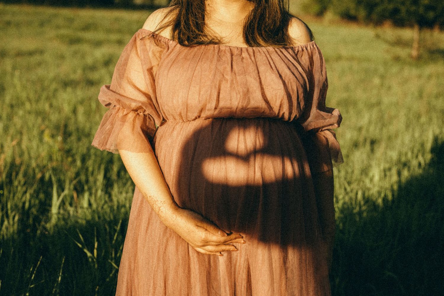 Pregnant woman in blush off-shoulder dress cradling baby bump with heart shadow in sunlit green field.