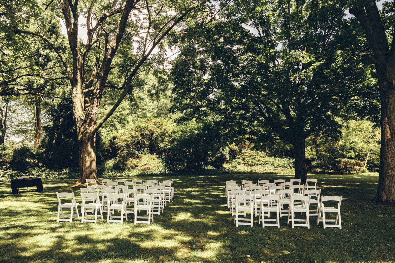 Outdoor wedding ceremony setup with white folding chairs arranged on green lawn under large trees.