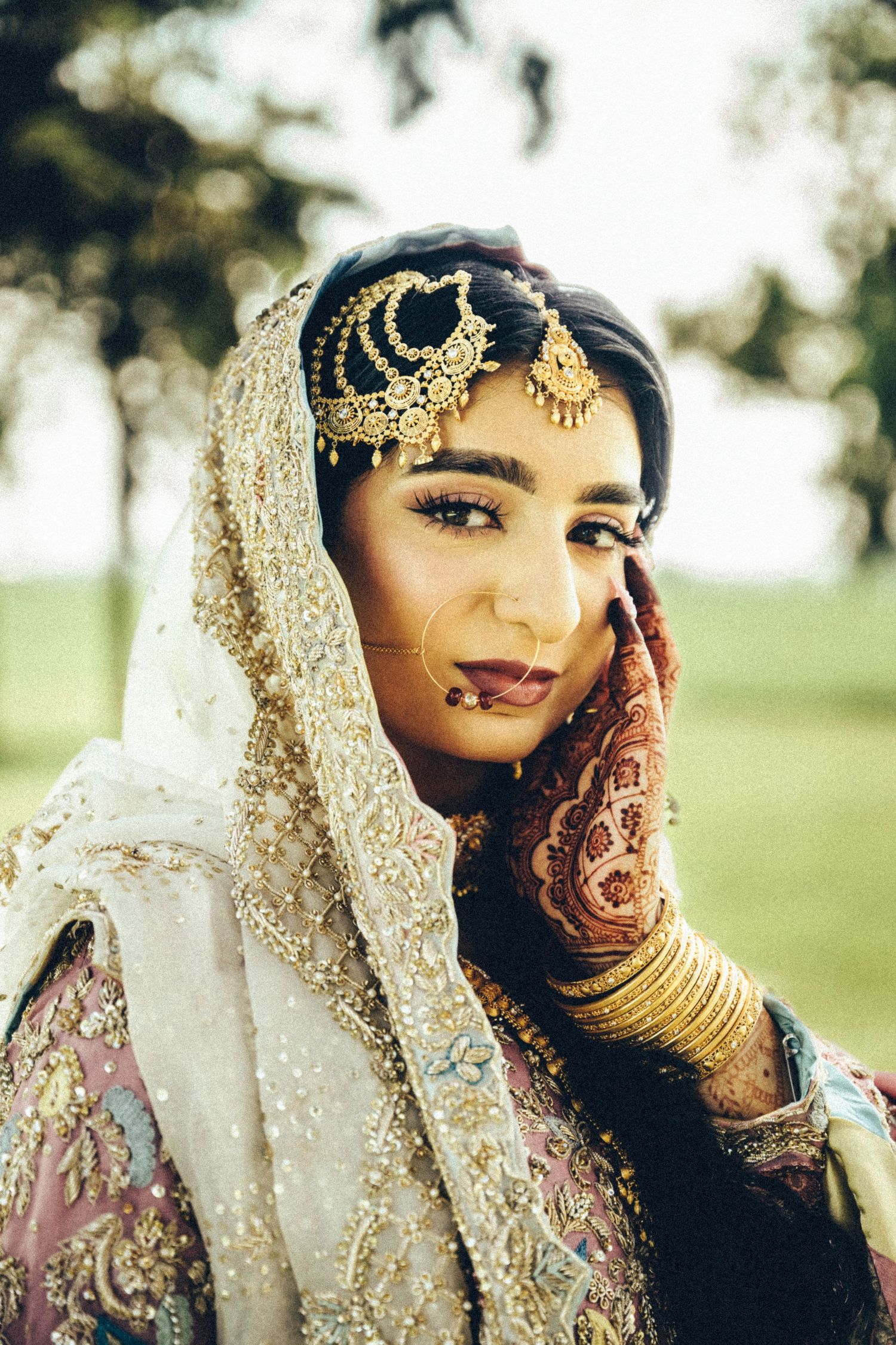 Stunning South Asian bride in gold maang tikka and embroidered dupatta with mehndi hands poses for a portrait outdoors.