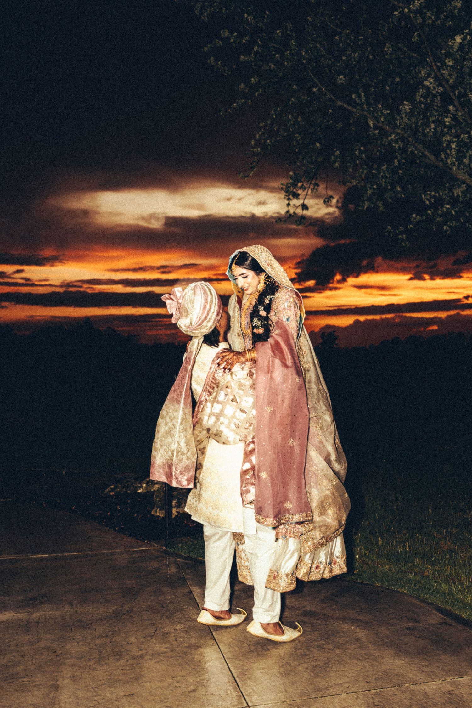 South Asian bride in pink lehenga embraces groom in ivory sherwani under a dramatic sunset sky.