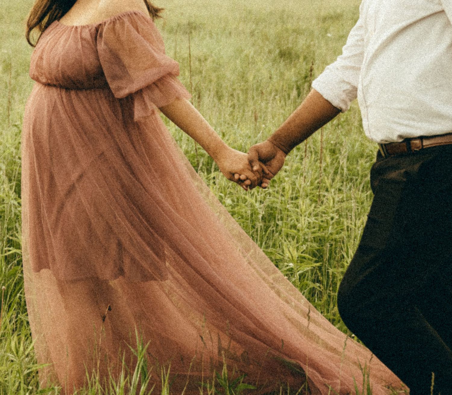 Pregnant woman in flowing dusty rose tulle gown holding hands with partner walking through green meadow.