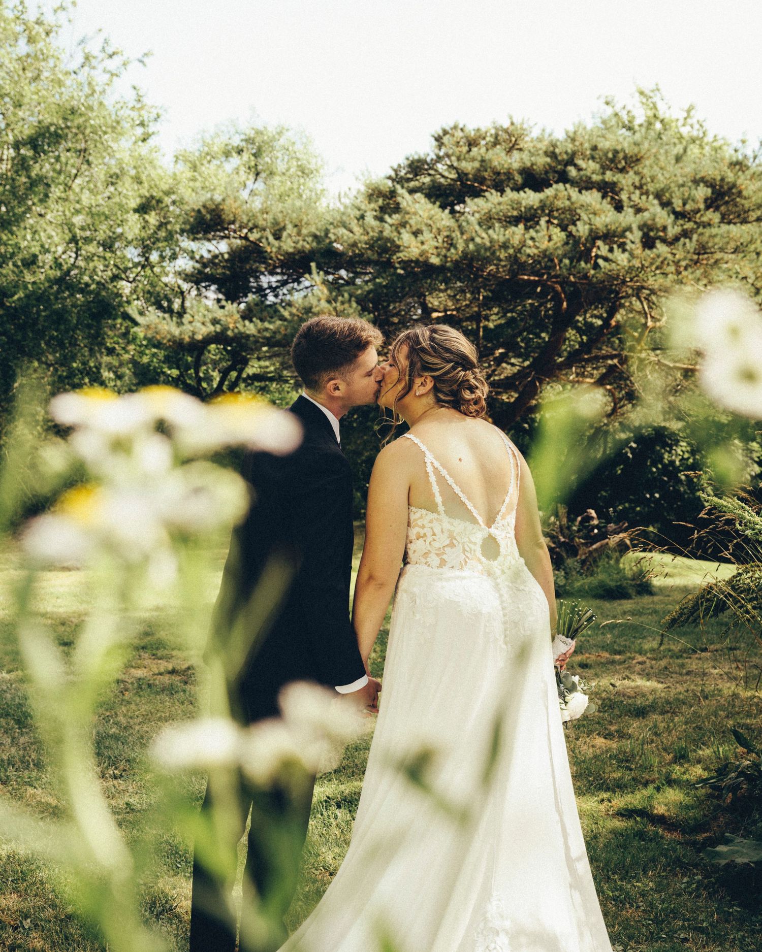 Groom kisses bride on the cheek as she holds bouquet, framed by white wildflowers and trees.