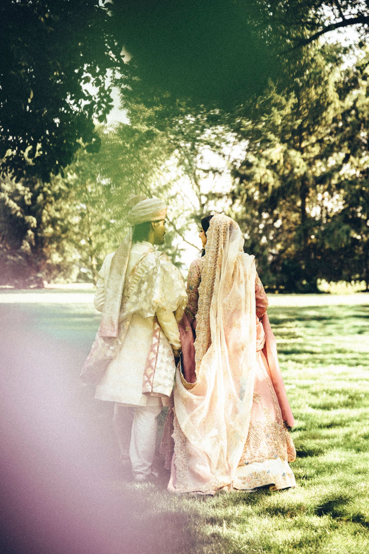South Asian bride and groom in traditional attire holding hands while walking through lush green park.