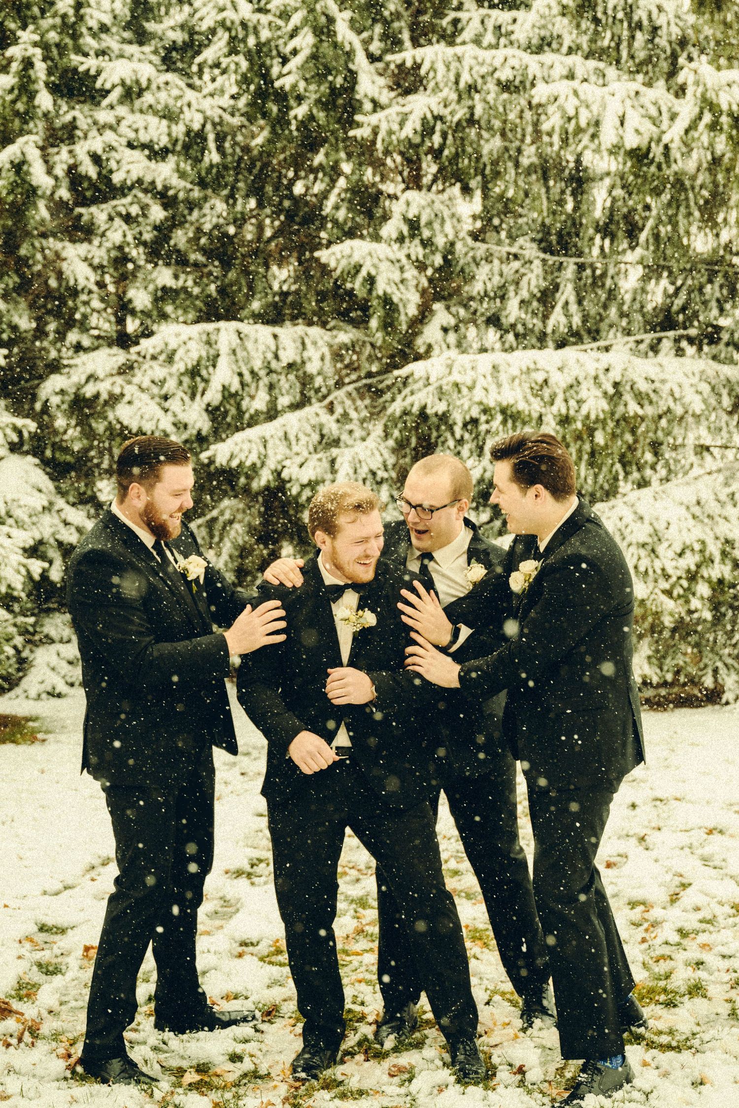 Four groomsmen in black suits laughing together in snowy winter wedding photo outdoors.