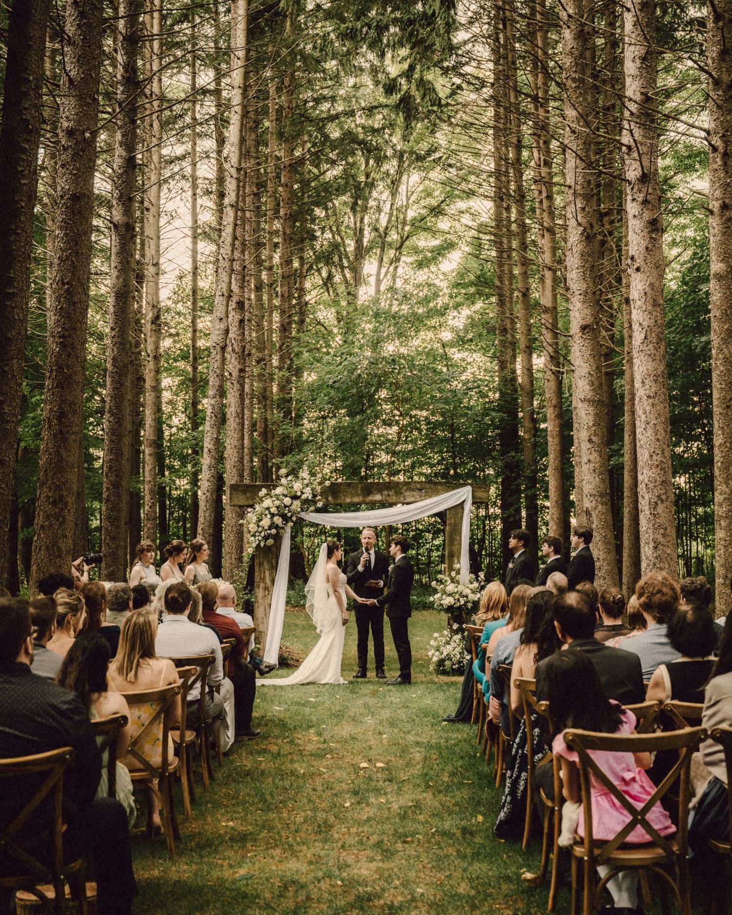 Romantic outdoor forest wedding ceremony with couples at floral arch aisle surrounded by tall trees and seated guests.