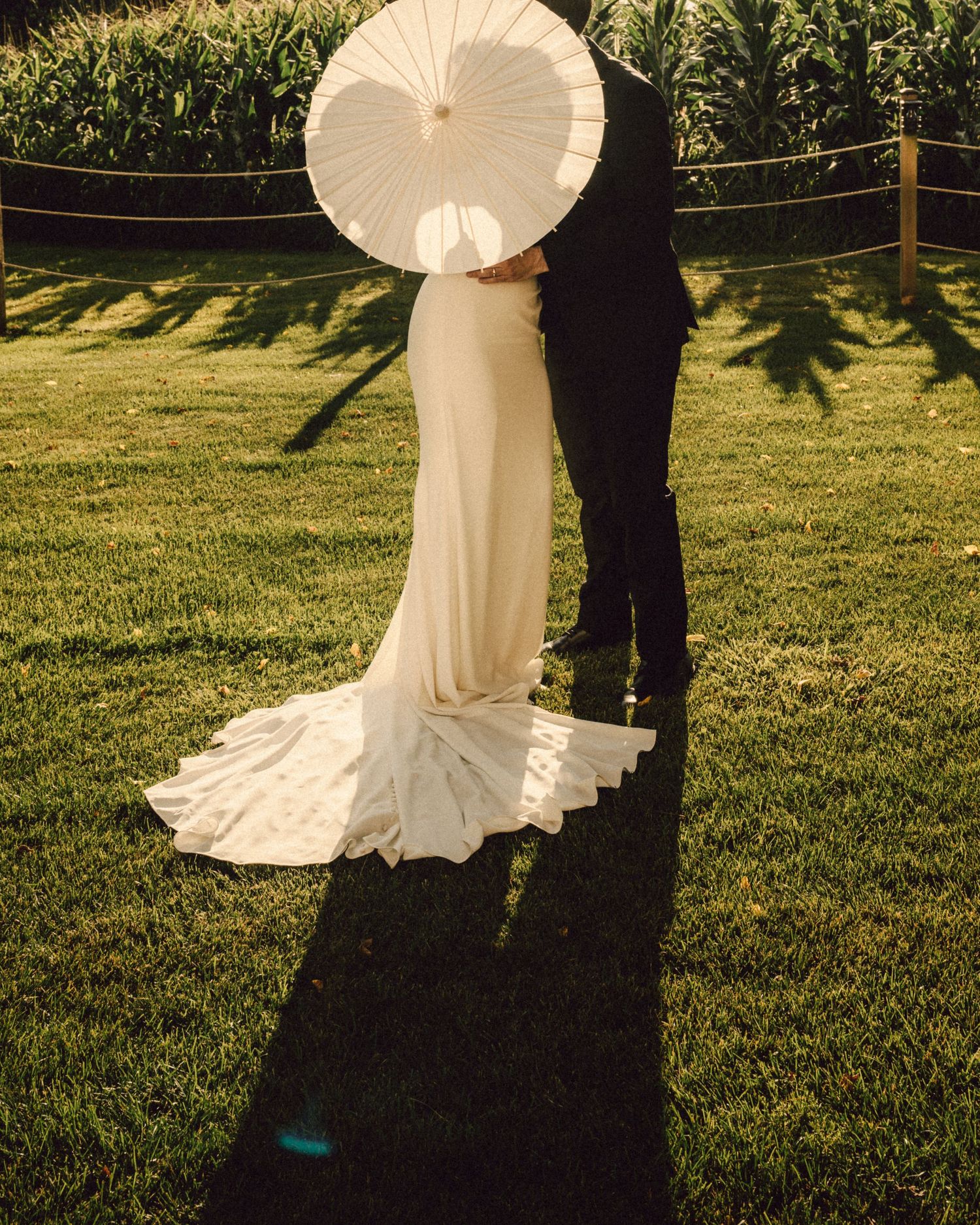 Bride and groom share a kiss hidden behind a white parasol casting shadows on green lawn.