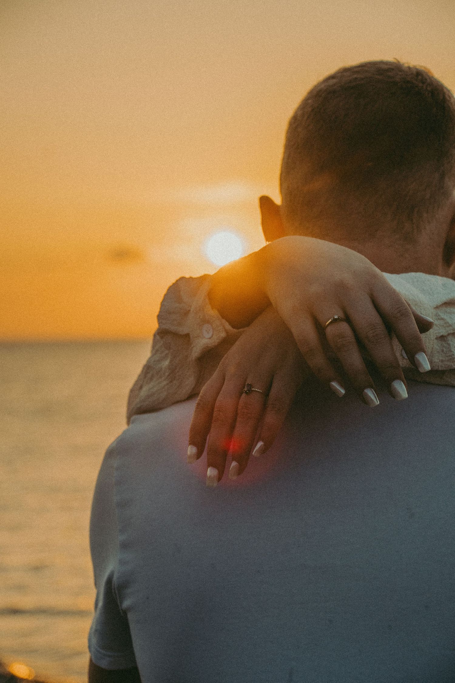 Couple embracing at golden sunset by the ocean, woman's hand with ring resting on man's shoulder.