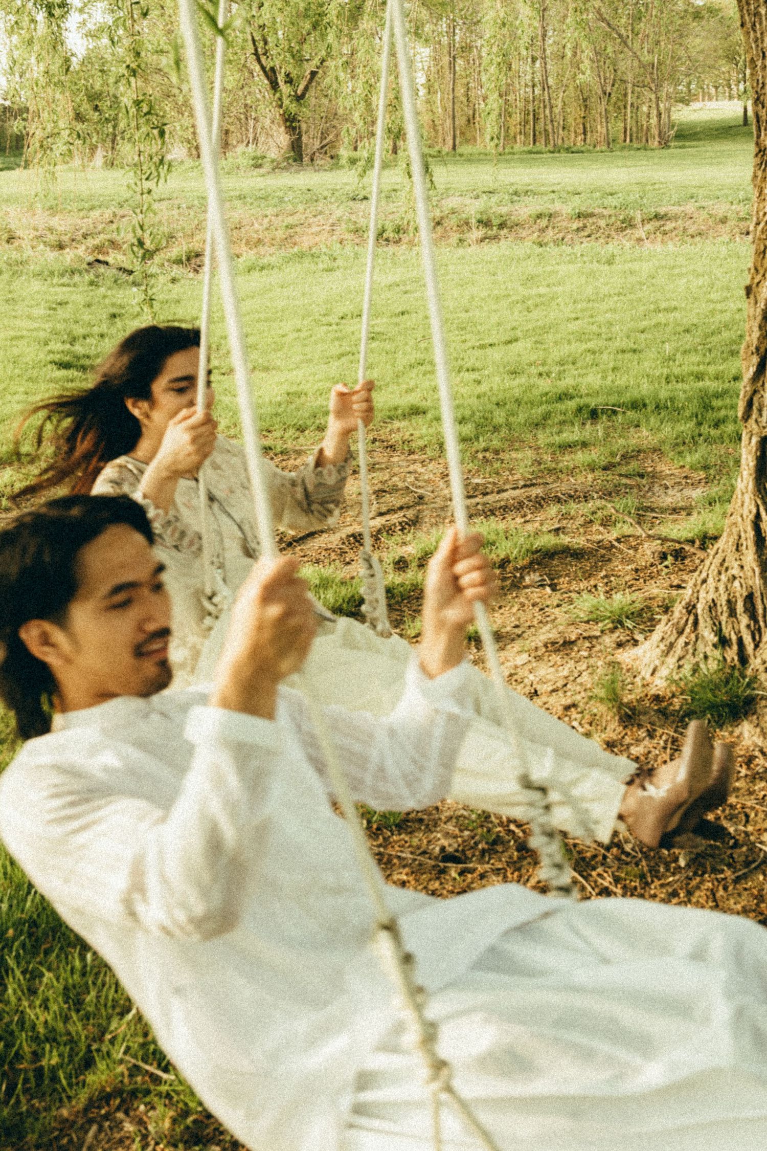 Couple in white outfits laughing on a rope swing in a sunny park.
