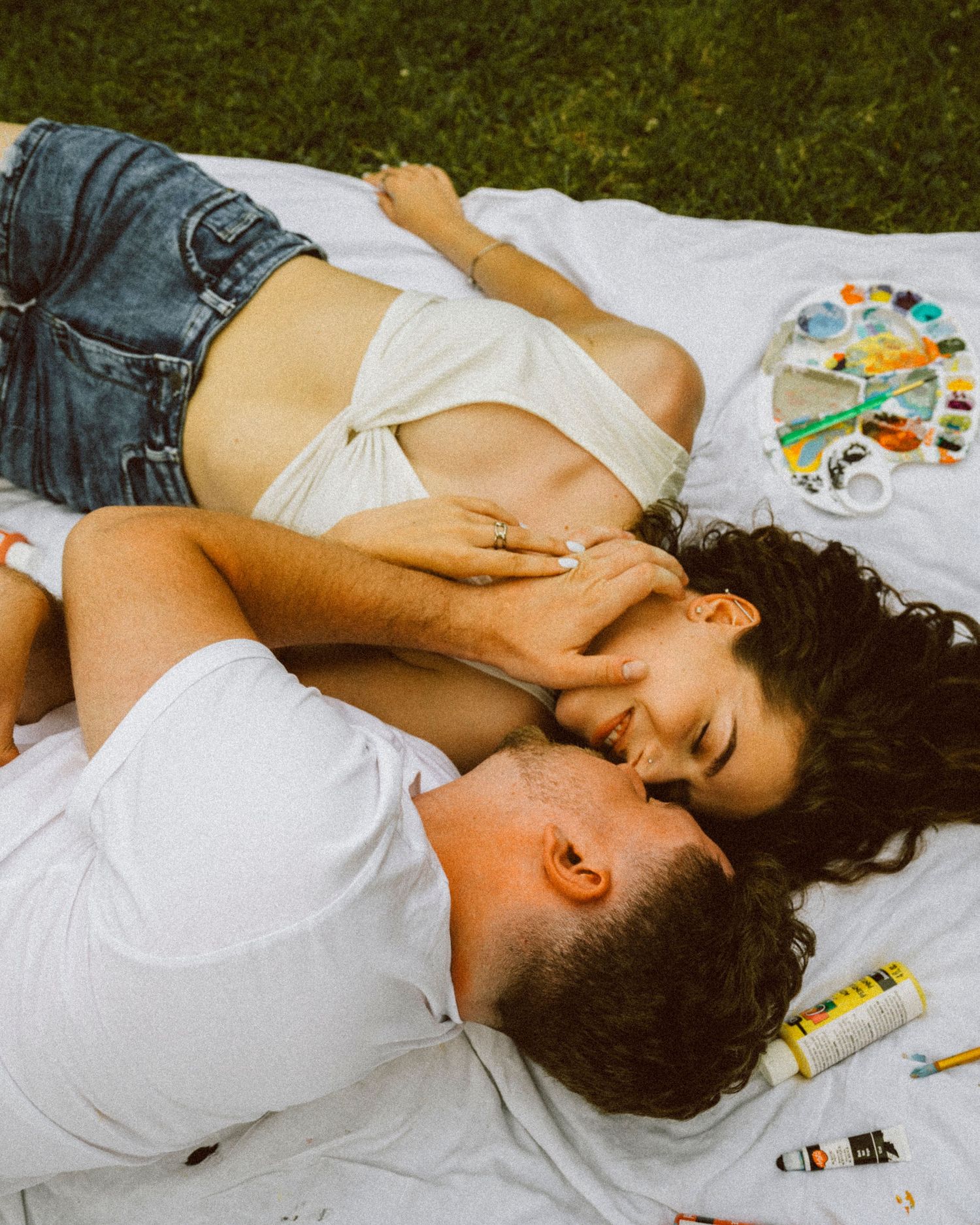 Couple lying on blanket laughing together surrounded by watercolor paints and art supplies.