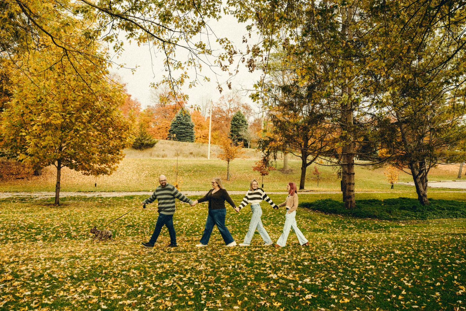 Family of four holding hands walking through park with golden autumn leaves on ground.