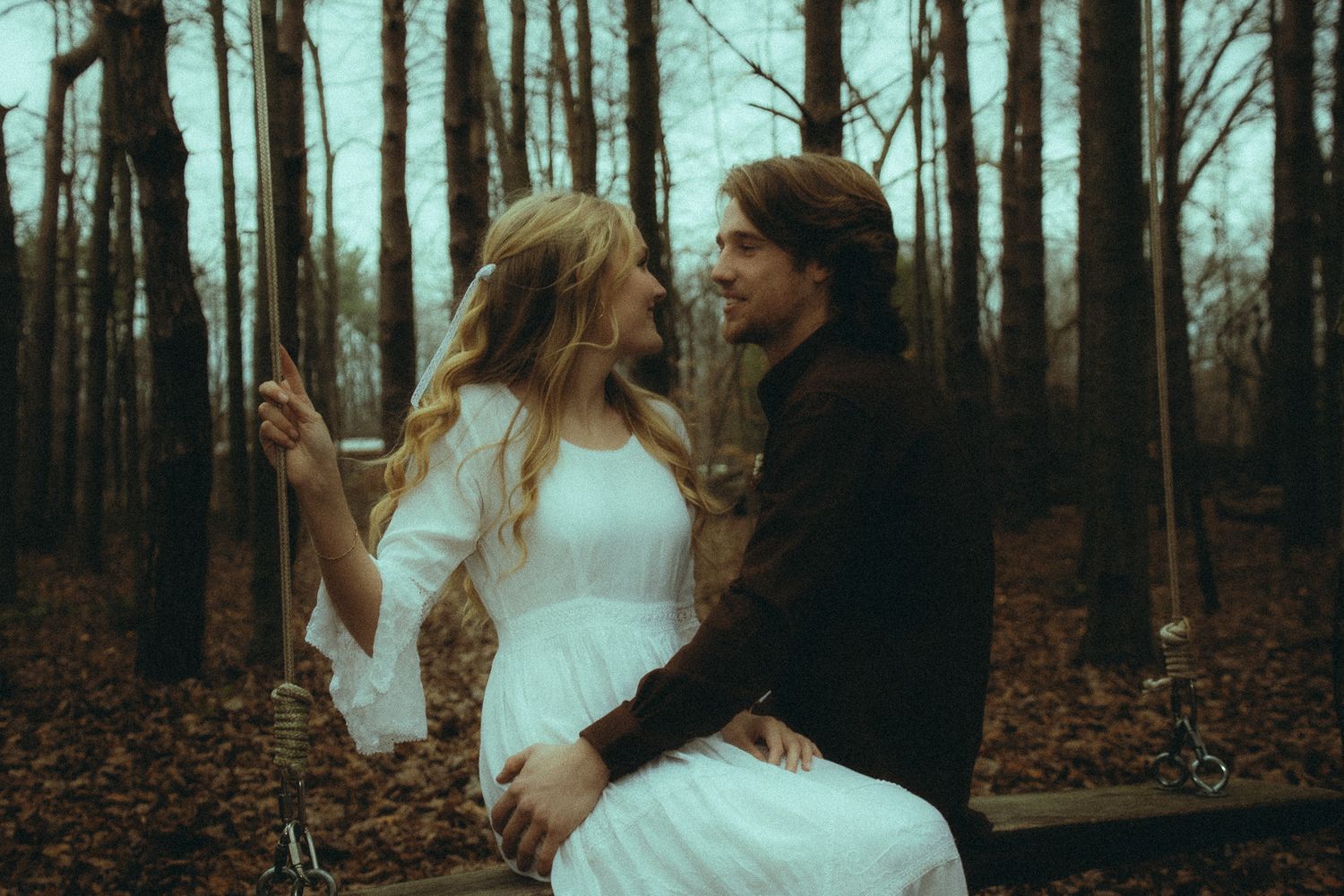 Romantic couple sitting on a swing in an autumn forest, smiling at each other in moody lighting.