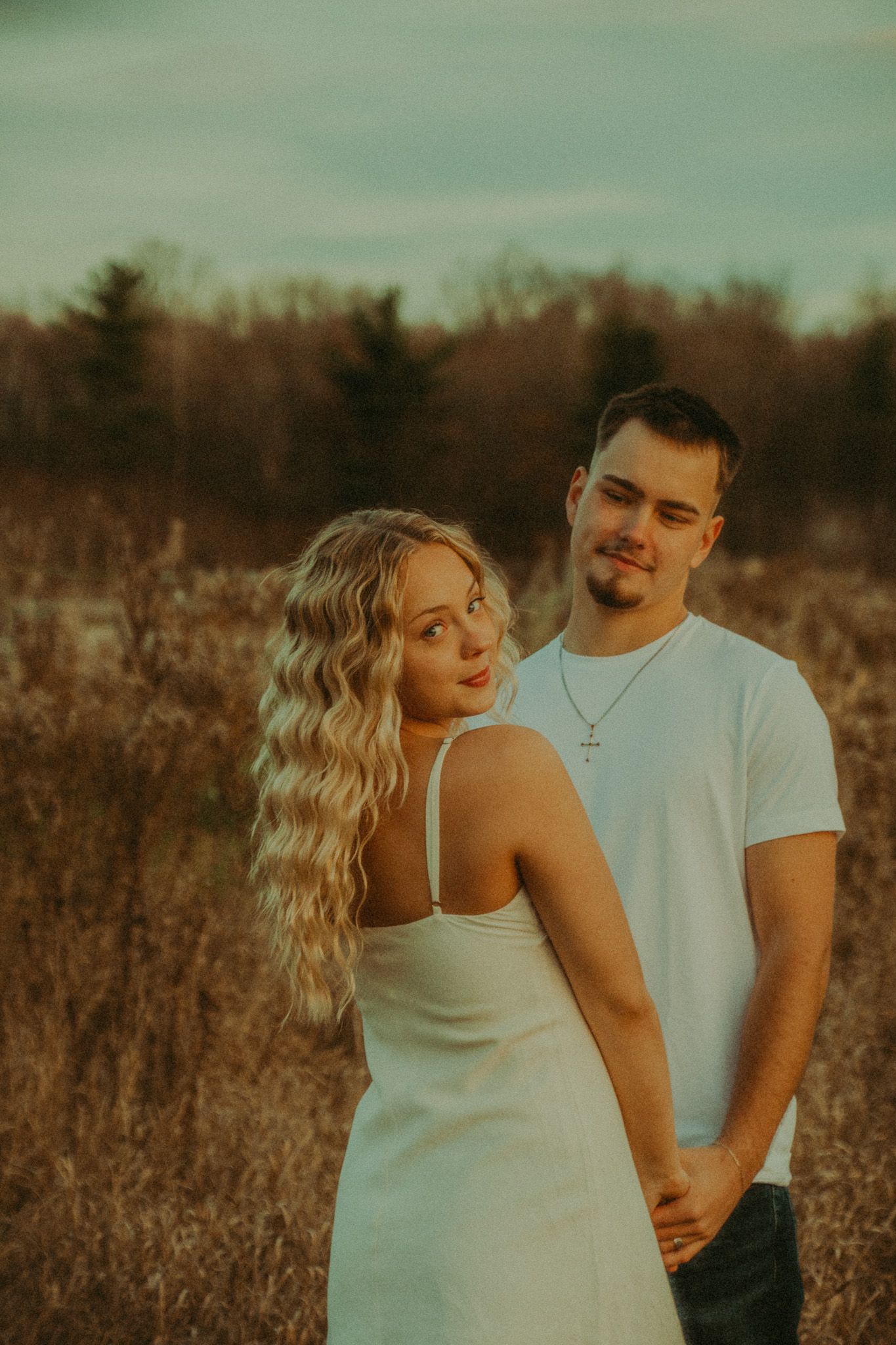Blonde woman in white dress looking back at camera while man in white tee stands behind her.