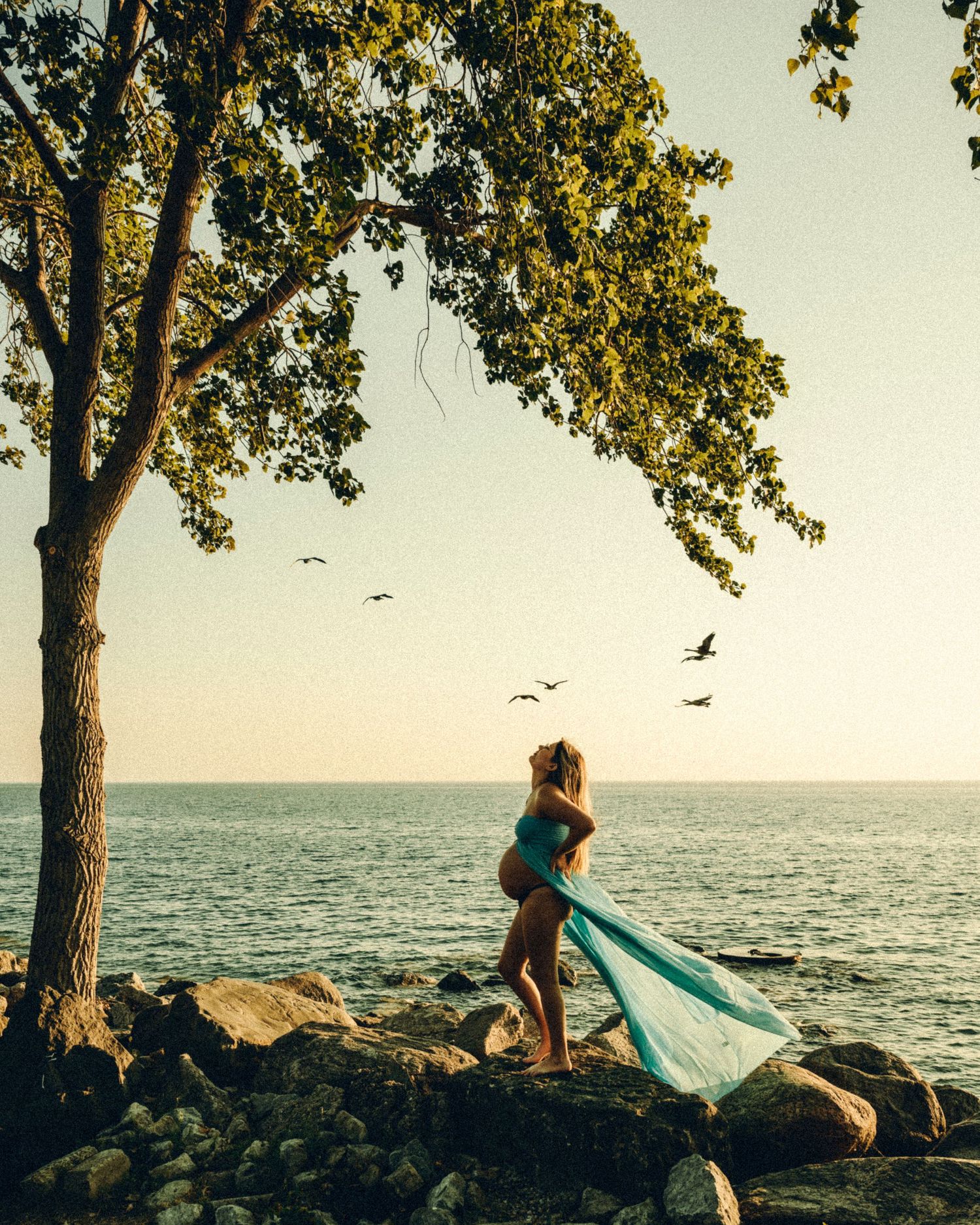 Pregnant woman in flowing teal dress standing on rocks by serene lakeside at sunset.