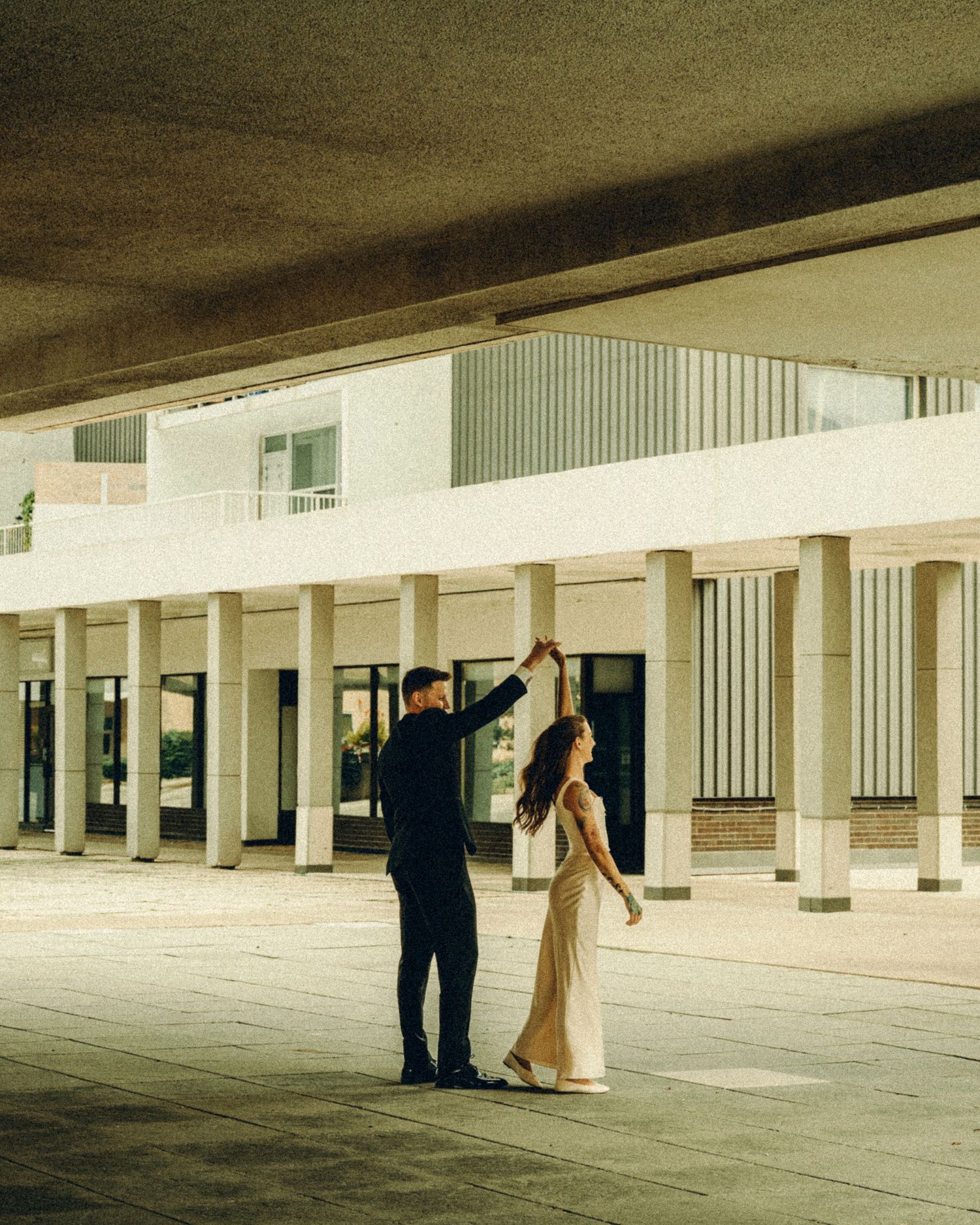 Bride and groom dancing elegantly under concrete colonnade in stylish wedding attire.