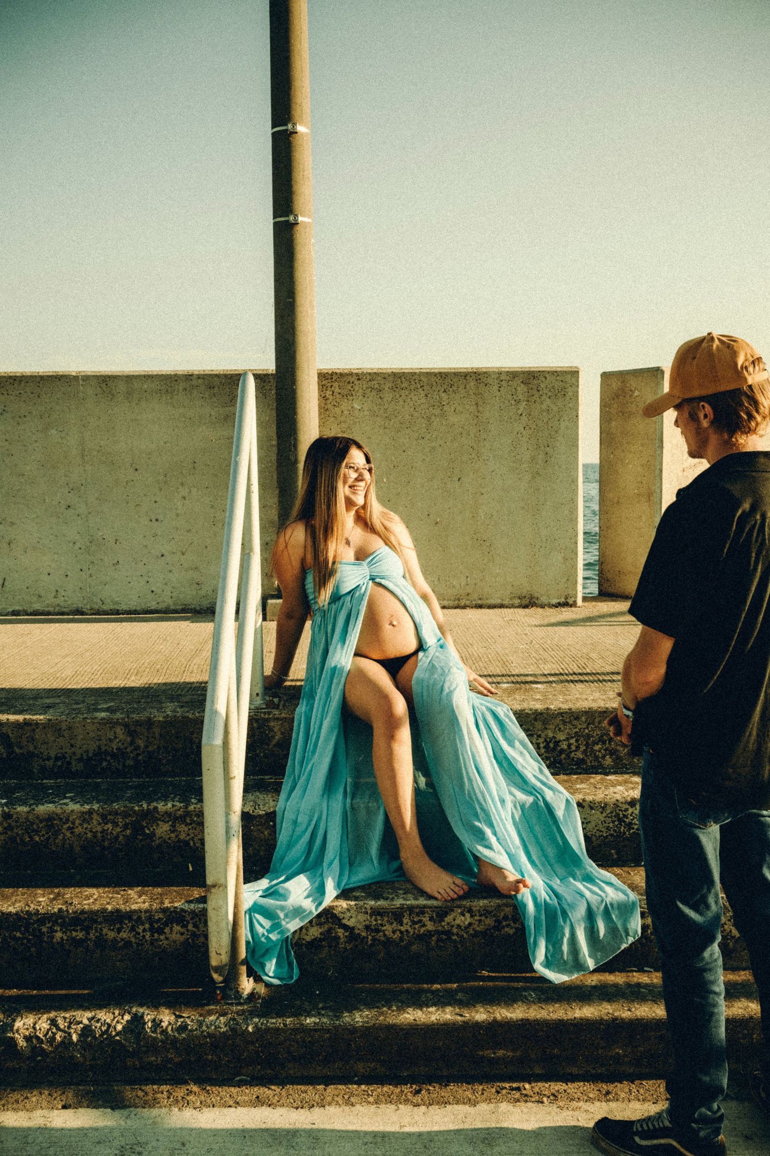 Pregnant woman in flowing teal maternity gown sits on steps smiling at her partner in golden sunset light.