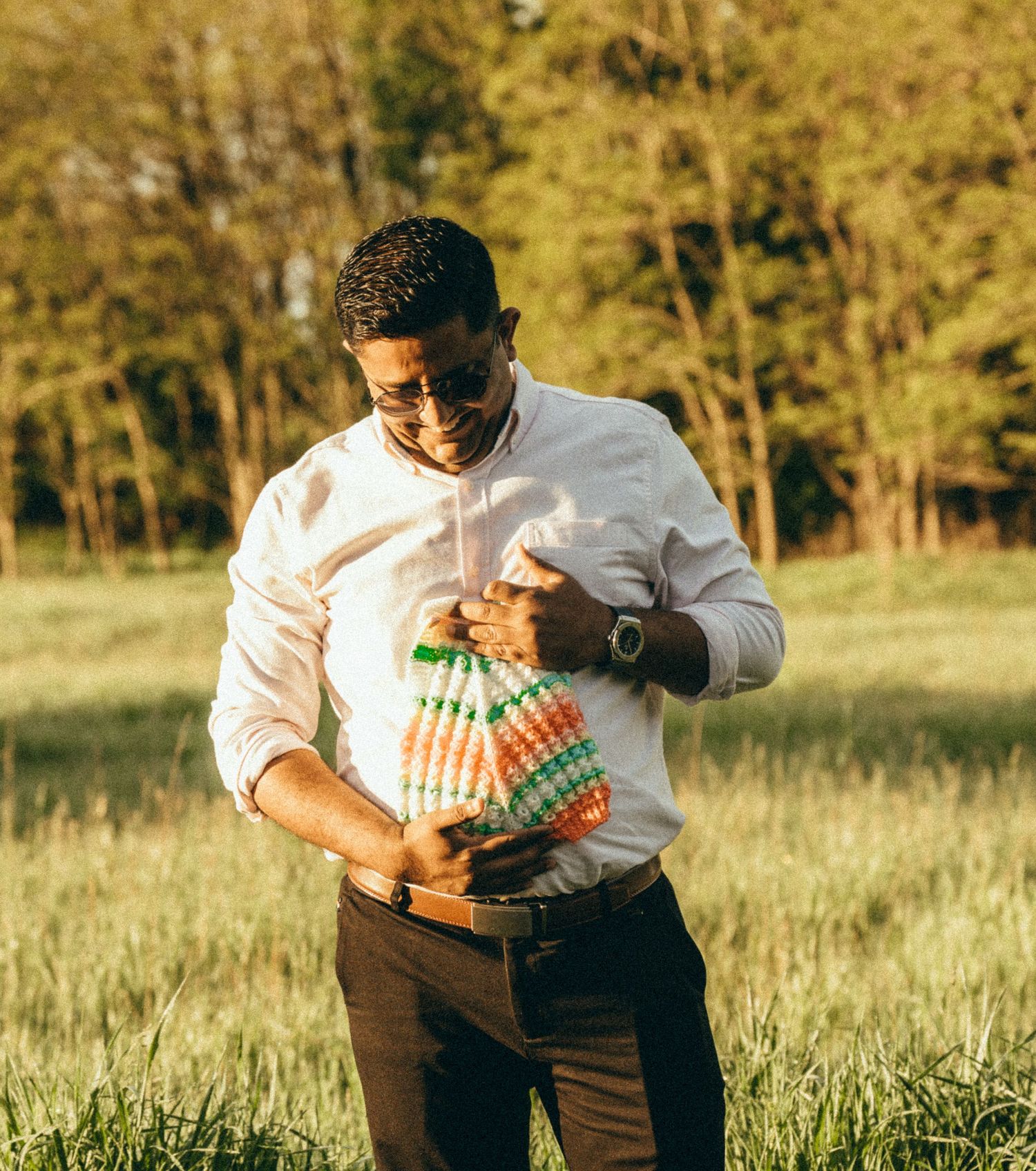 Man in white shirt holding a newborn baby wrapped in colorful blanket outdoors in a sunny green field.