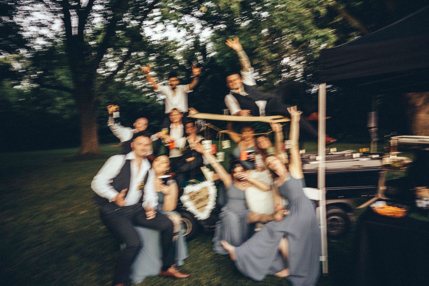 Wedding party celebrates joyfully on vintage vehicle surrounded by trees at outdoor evening reception.