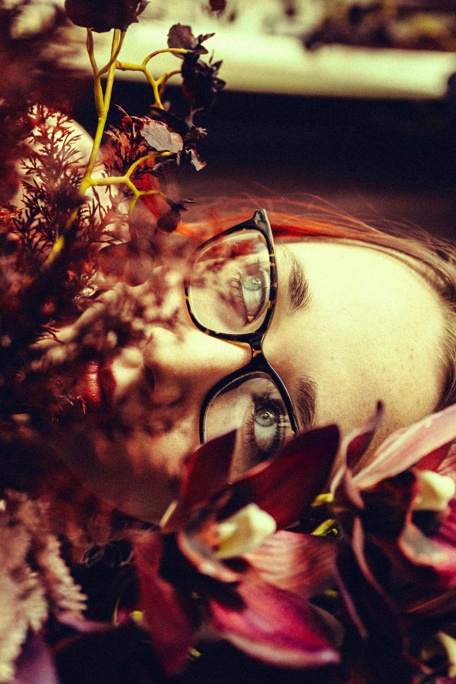 Woman with red hair and glasses partially hidden among dark red and purple flowers.