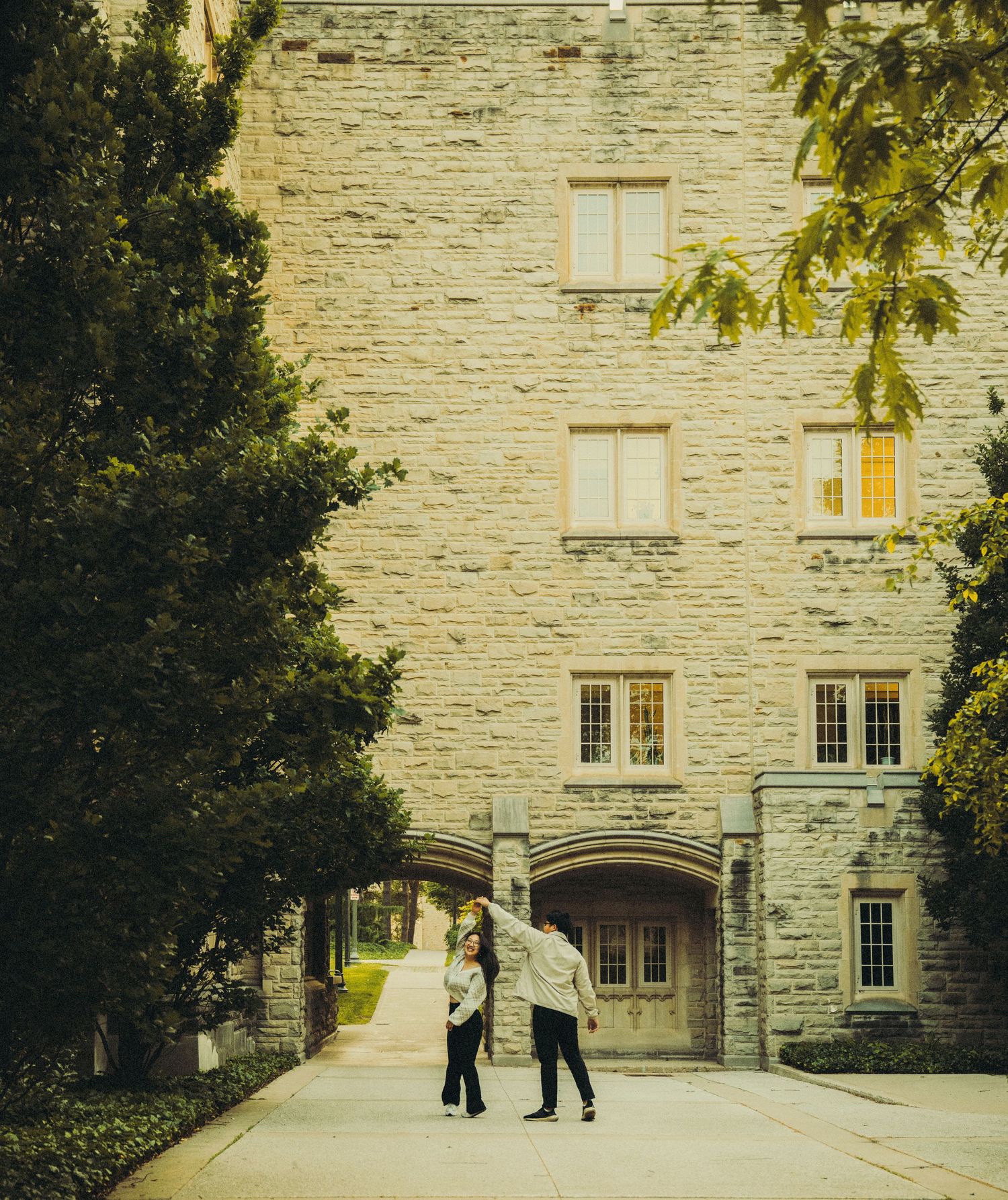 Couple standing together outside a historic stone university building surrounded by lush green trees.
