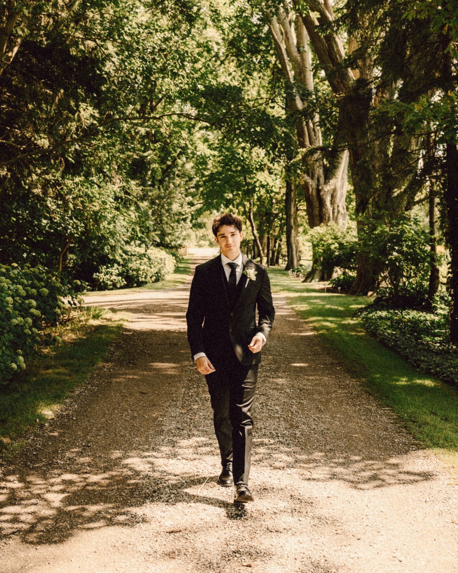Dapper groom in black suit walks confidently down a tree-lined gravel path on his wedding day.
