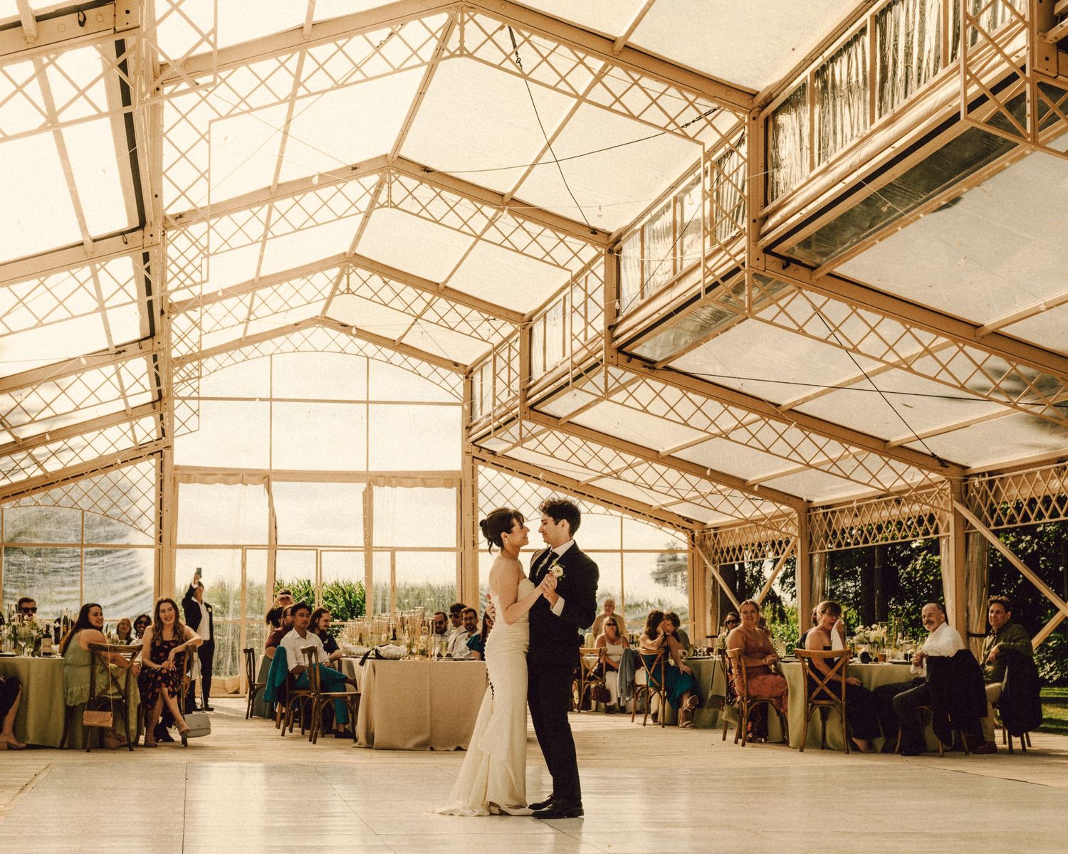 Couple shares first dance in elegant glass greenhouse wedding venue with guests seated at decorated tables.