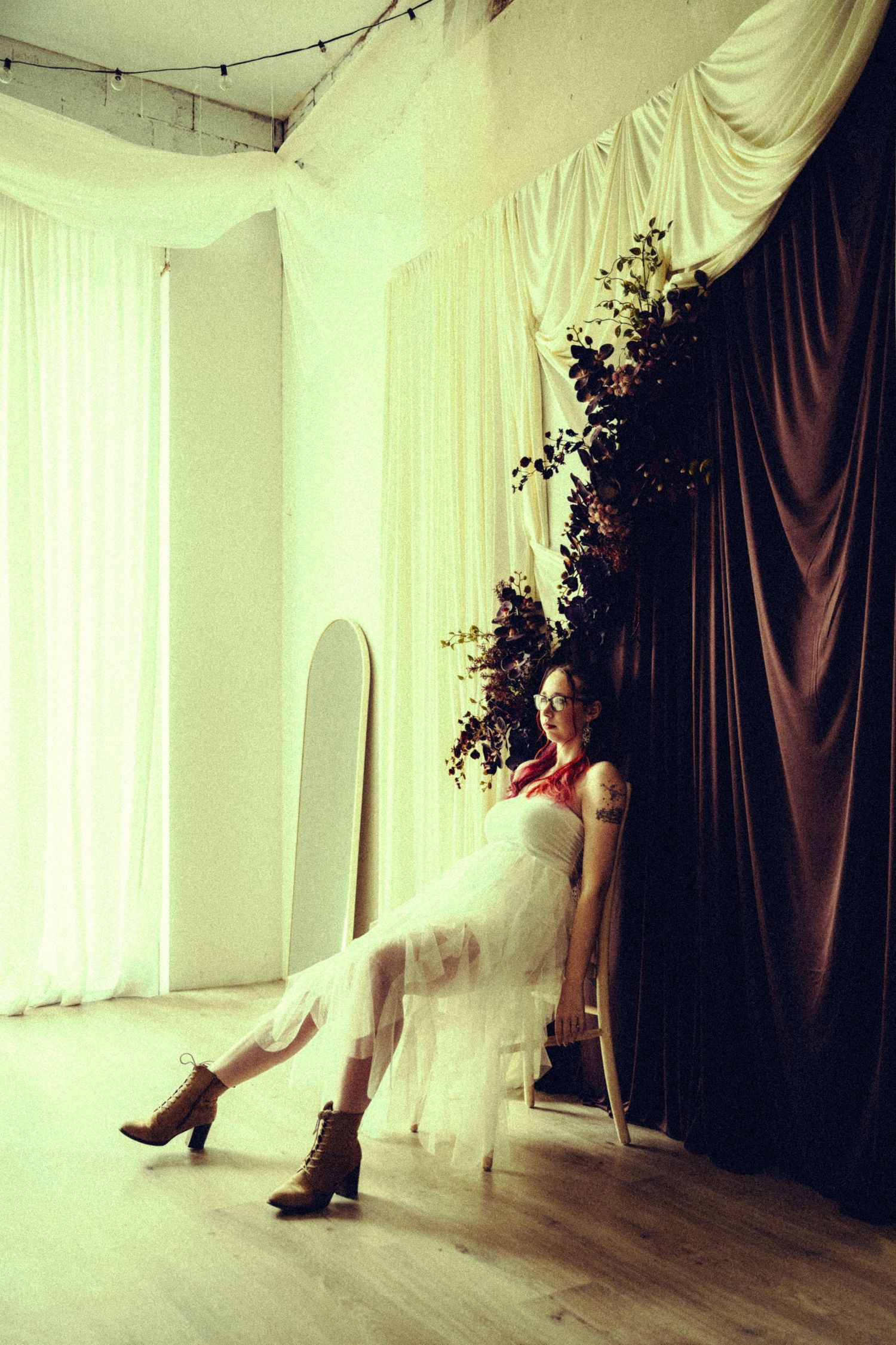 Woman in white tulle dress and boots sitting in chair beside dark draped curtain indoors.