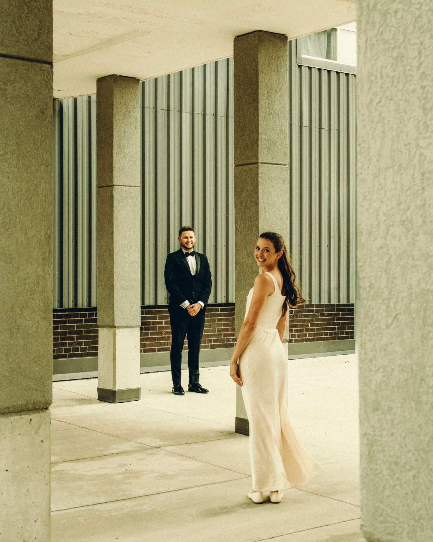 Bride in white gown smiling at camera while groom stands in background between columns.