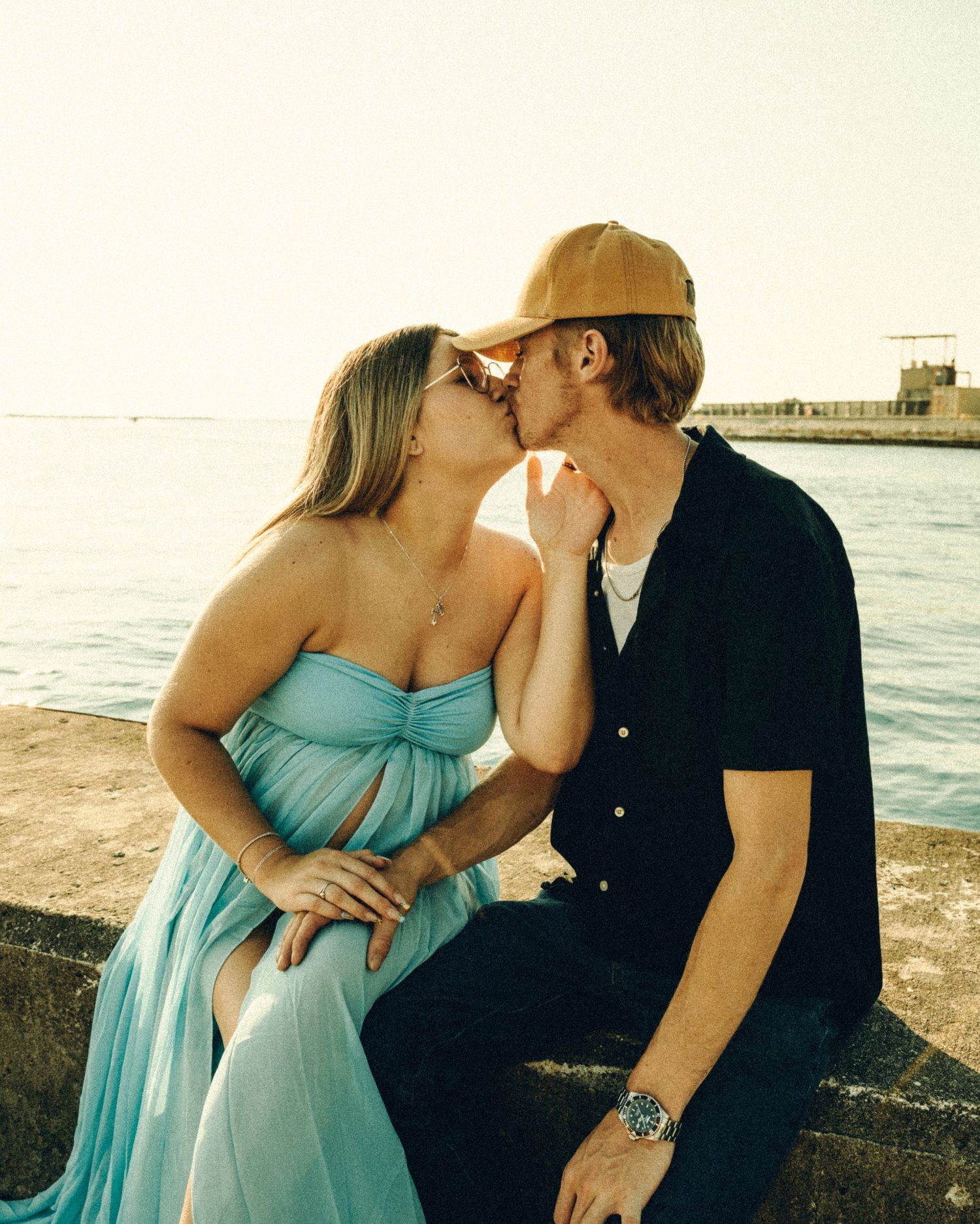 Couple sharing a tender kiss while sitting on a seaside wall, woman in blue dress and man in black shirt.