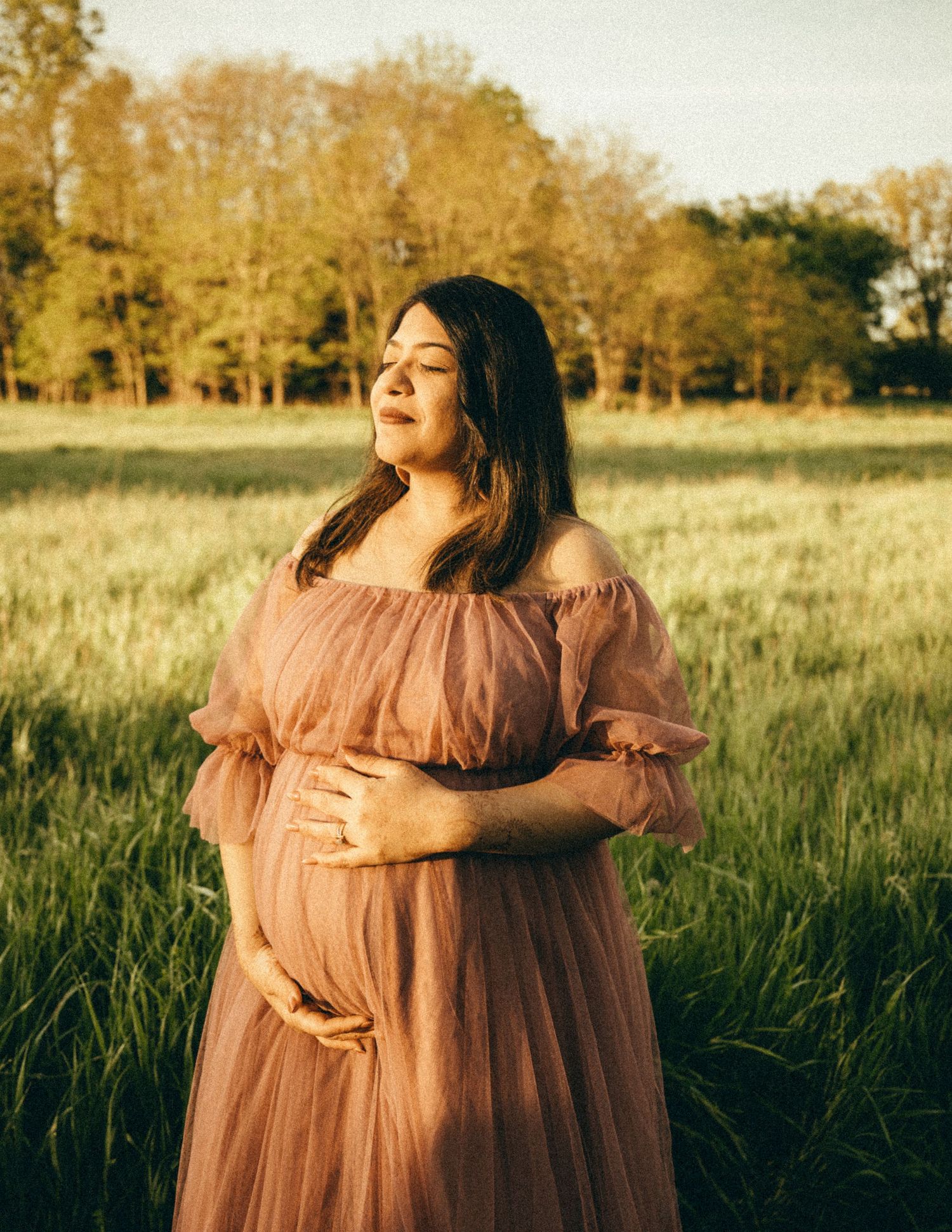 Glowing pregnant woman in blush off-shoulder tulle gown cradling her bump in a golden sunlit field.