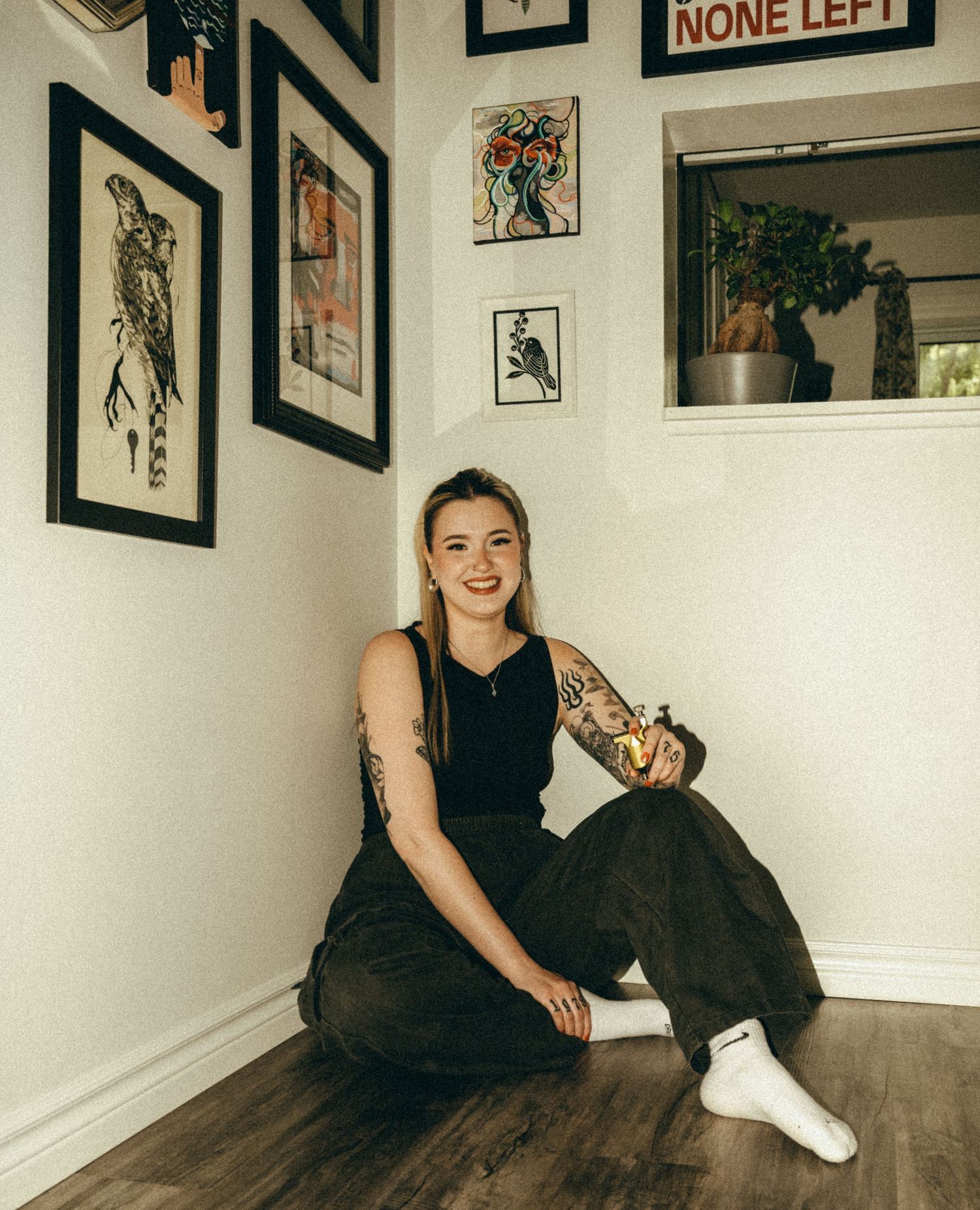 Smiling tattooed woman sitting on floor against white wall gallery of framed art prints in black pants and white socks.
