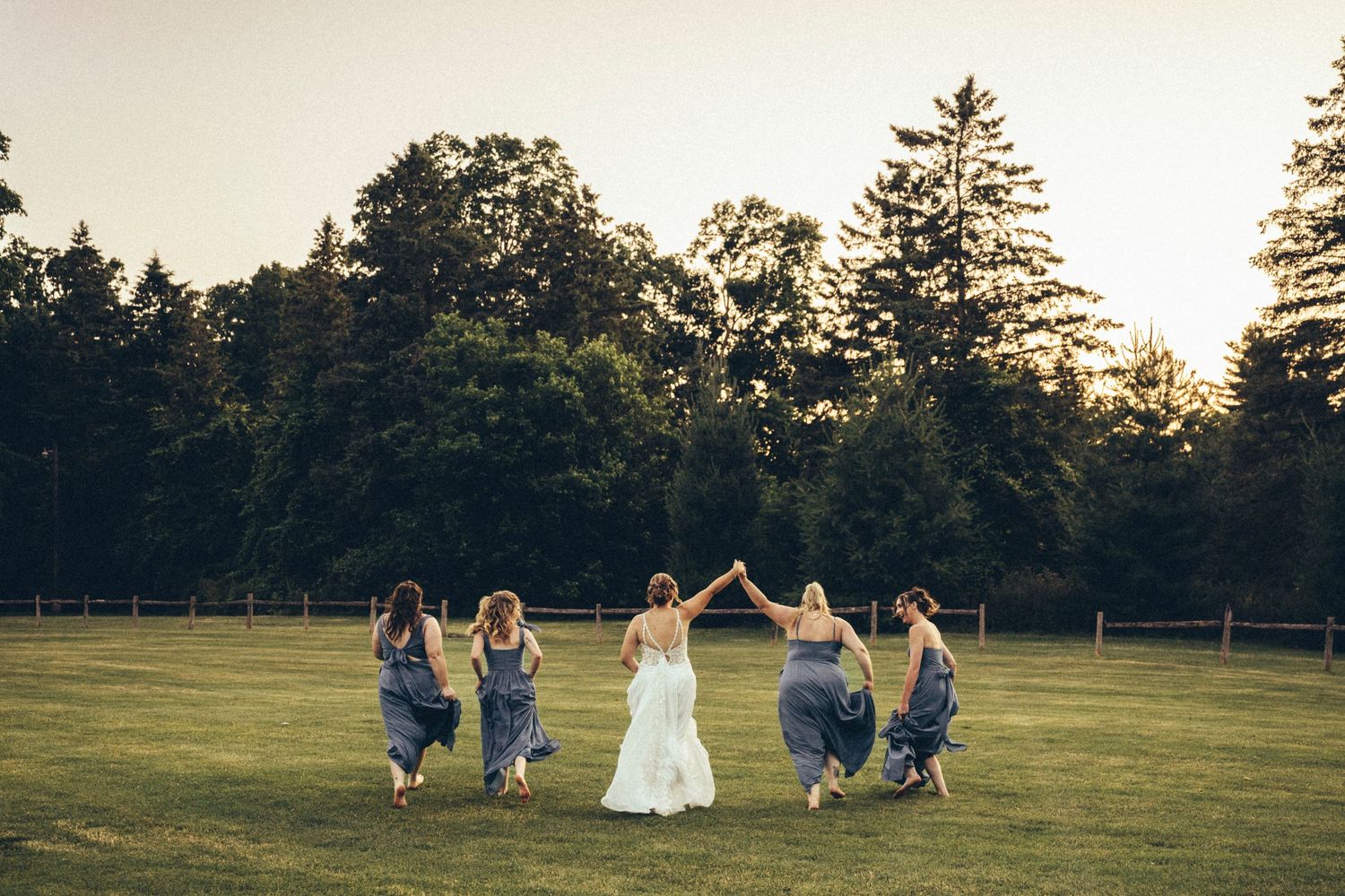 Bridesmaids in blue dresses hold hands with bride in white gown walking across green field at sunset.
