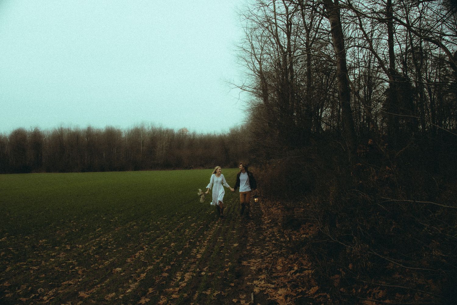 Two women in white dresses walking through a field at dusk near a treeline.