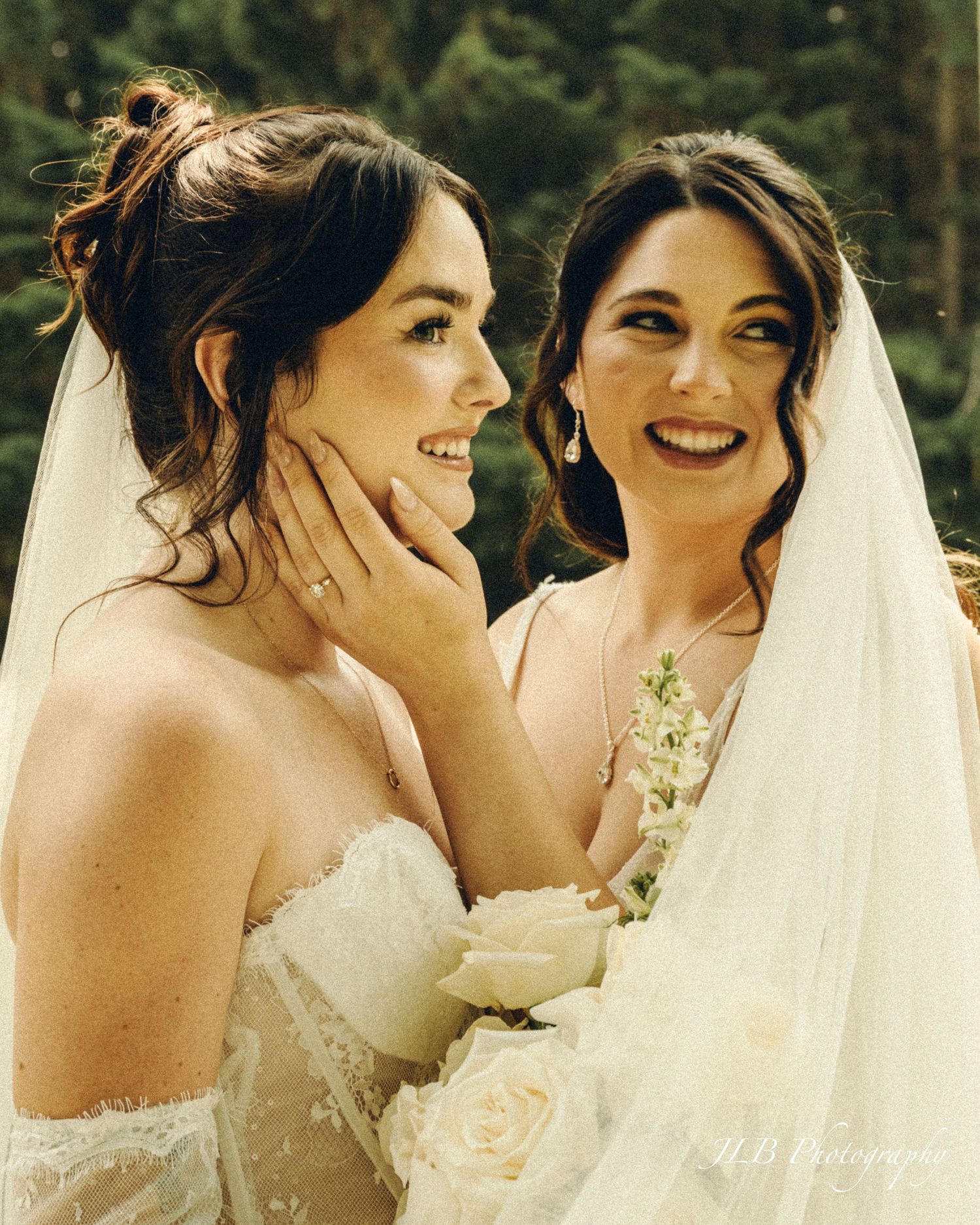 Two smiling brides in wedding dresses share a joyful moment, one tenderly touching the others face.